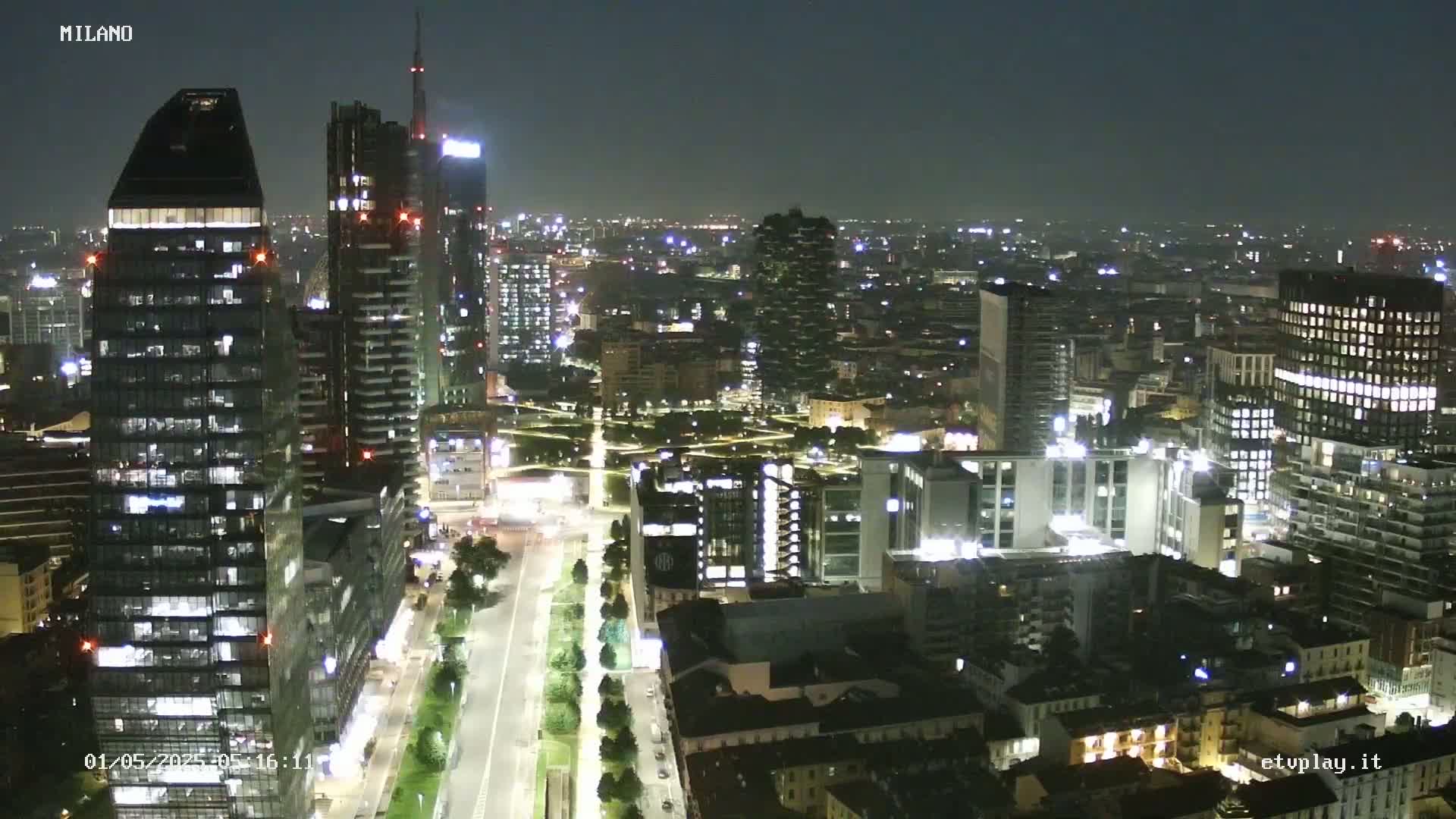A nighttime aerial view of a city with numerous illuminated skyscrapers and buildings, lit streets, and a park-like area.