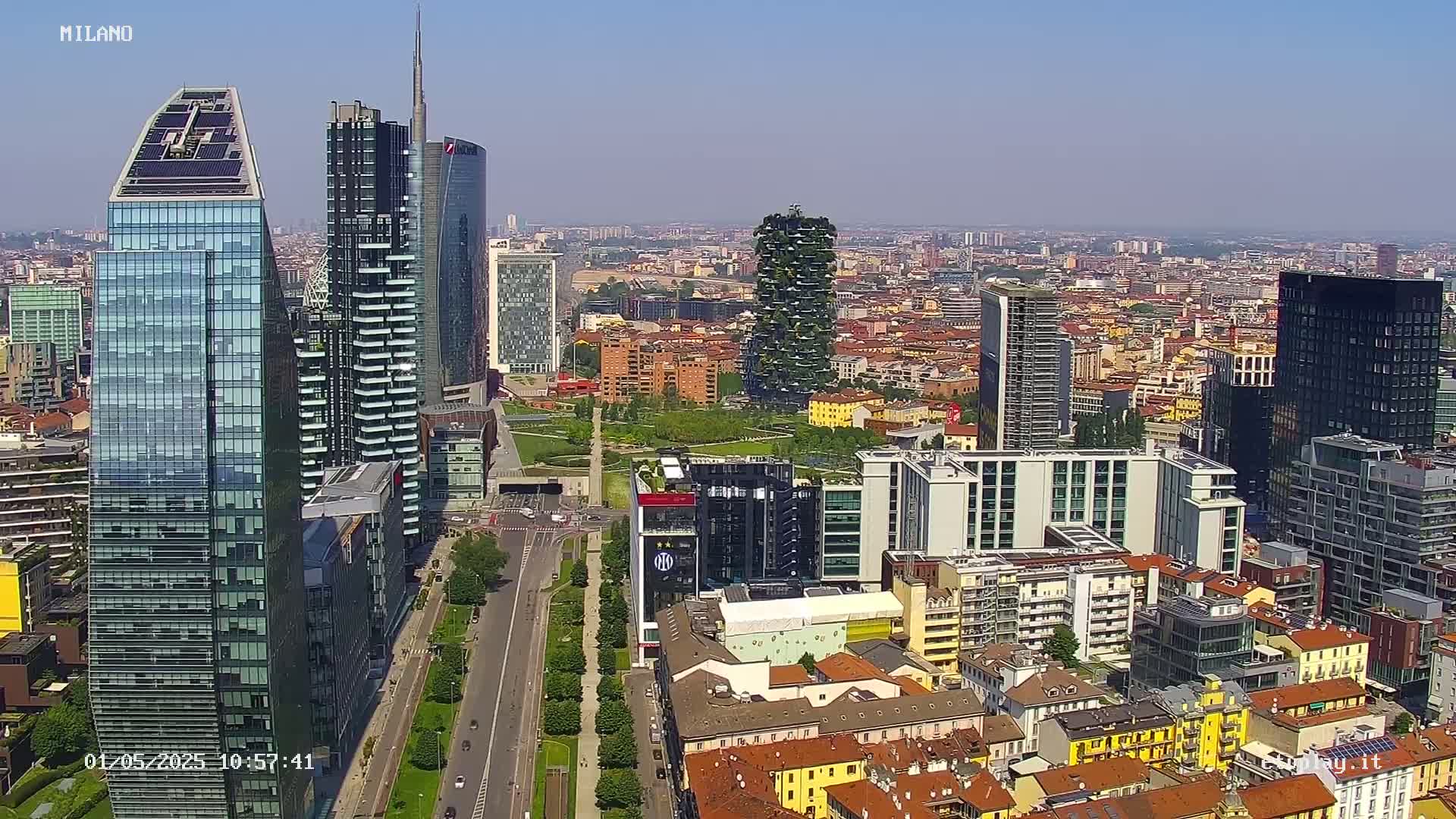 An aerial view of a city skyline on a clear, sunny day, showing numerous skyscrapers and surrounding buildings.