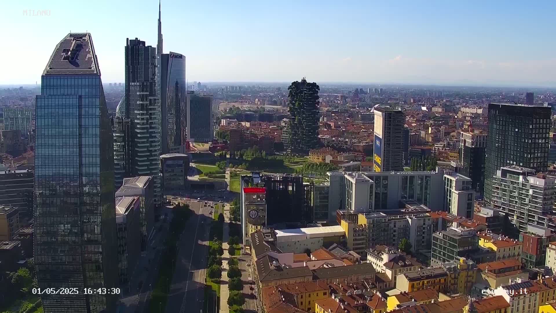 A high-angle, long shot of a city skyline on a sunny day, showing a mix of modern skyscrapers and traditional low-rise buildings.