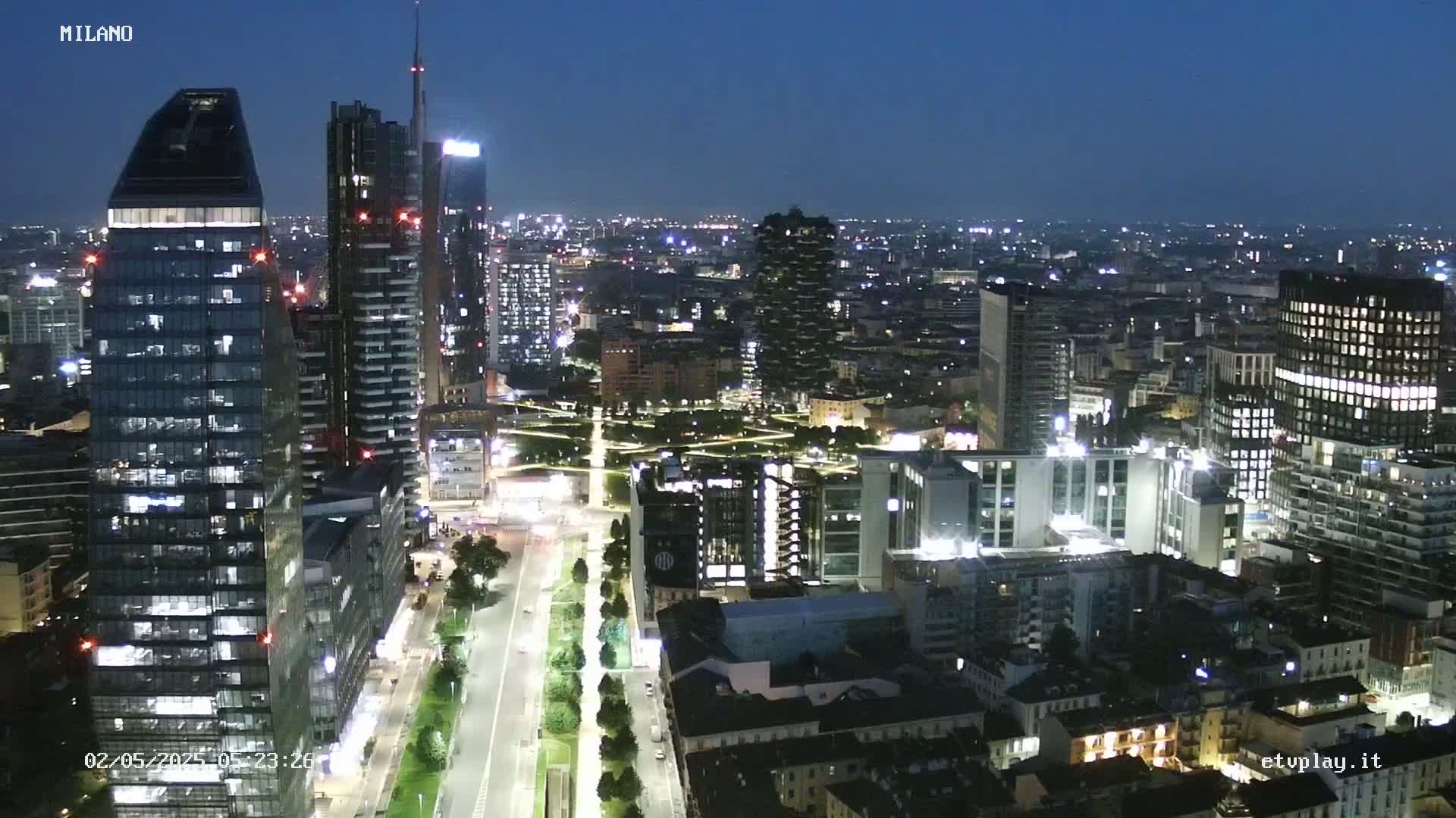 A nighttime aerial view of a city, showing numerous illuminated skyscrapers and buildings, with a long road and park visible below.