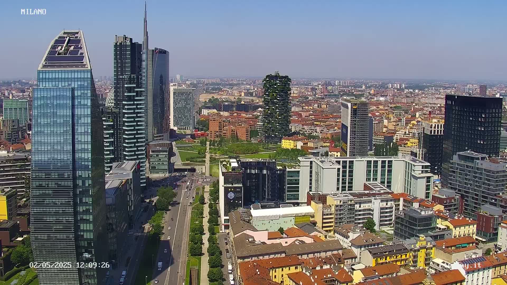An aerial view shows a modern city skyline under a clear, sunny sky, with tall glass skyscrapers and a mix of residential buildings surrounding a park.