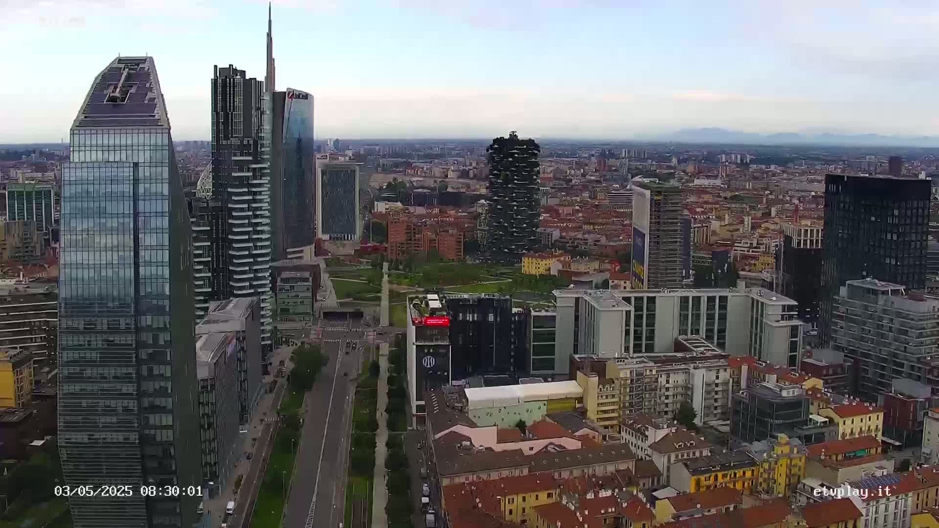 An aerial view shows a cityscape of numerous skyscrapers and buildings under a cloudy sky.