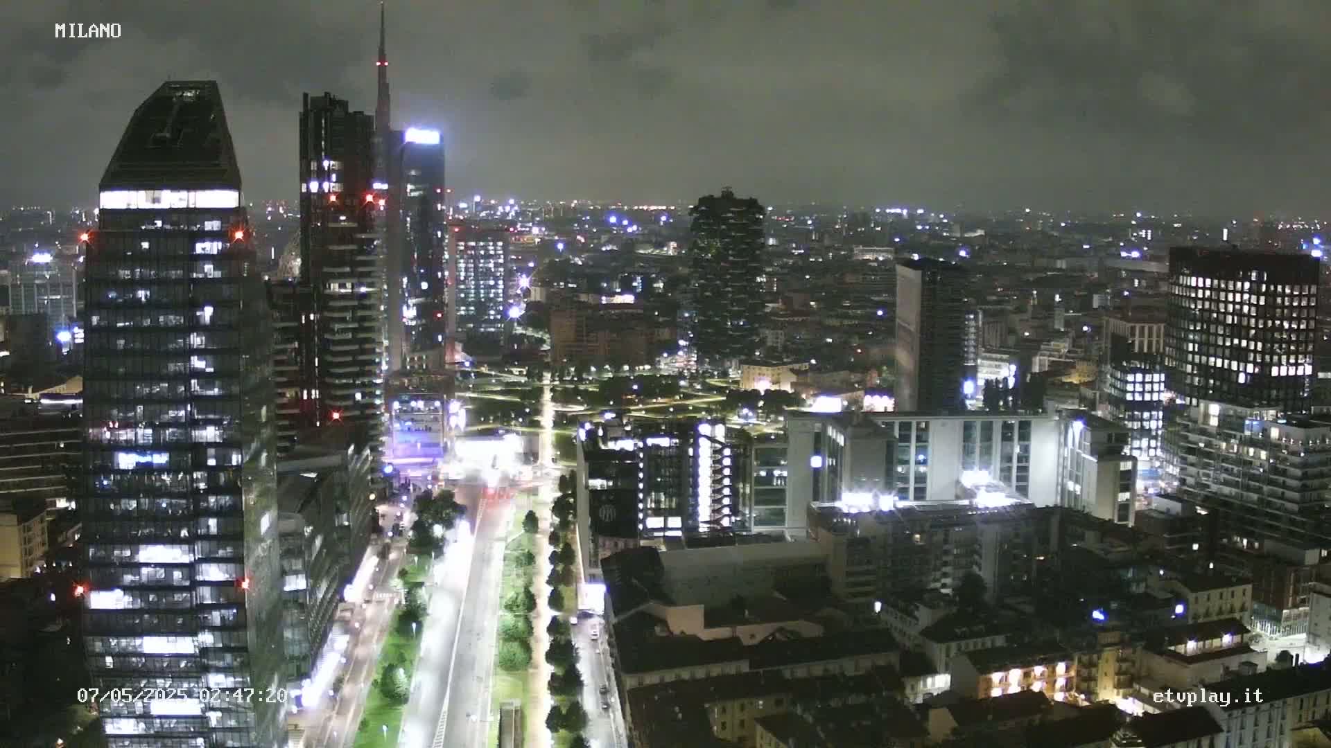 A nighttime aerial view of a city, showing numerous illuminated buildings and streets under a dark, cloudy sky.