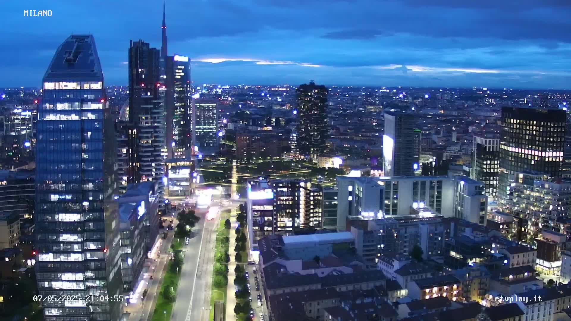 A nighttime aerial view of a city with numerous skyscrapers and illuminated streets under a partly cloudy blue sky.