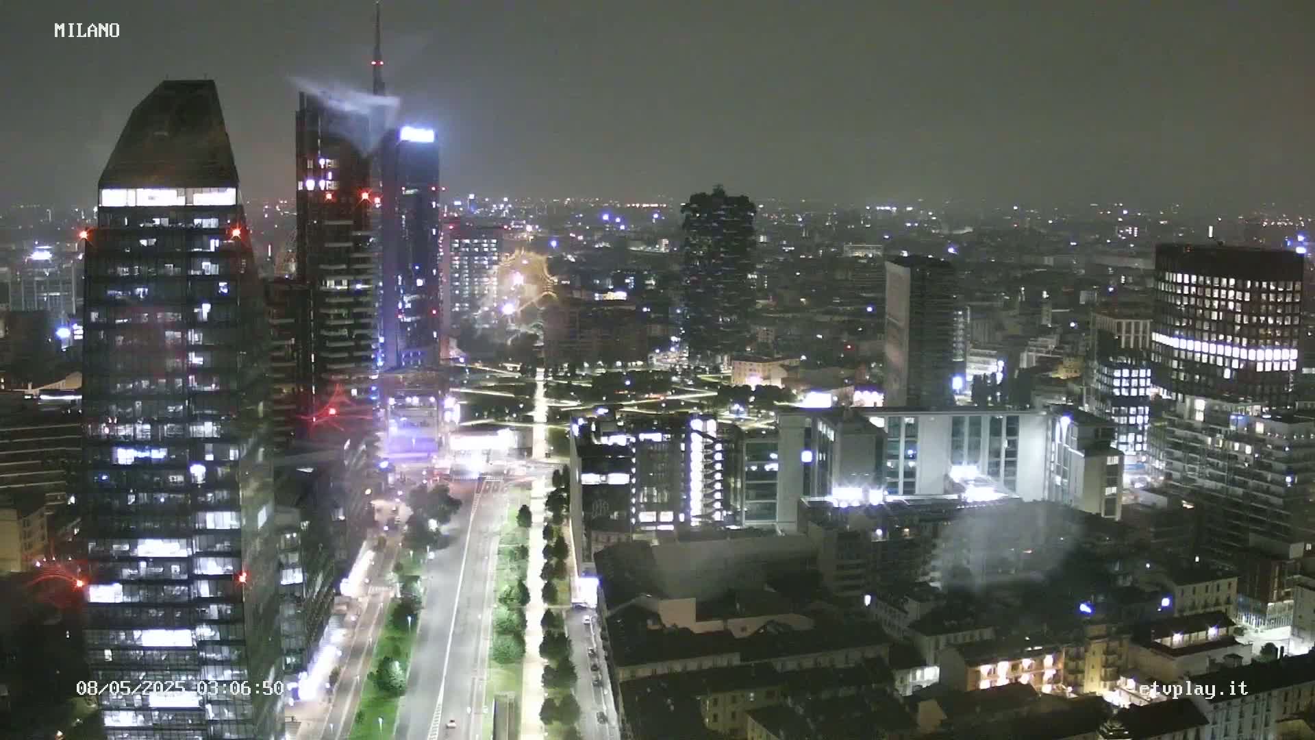 A nighttime, hazy view of a city skyline showing numerous illuminated skyscrapers and buildings along a wide road.