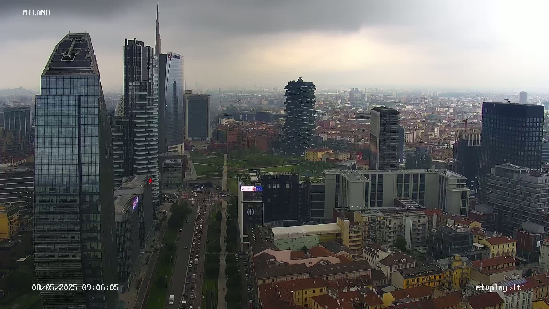 A high-angle, long shot of a city skyline on an overcast day shows numerous skyscrapers and surrounding buildings, with a road visible below.