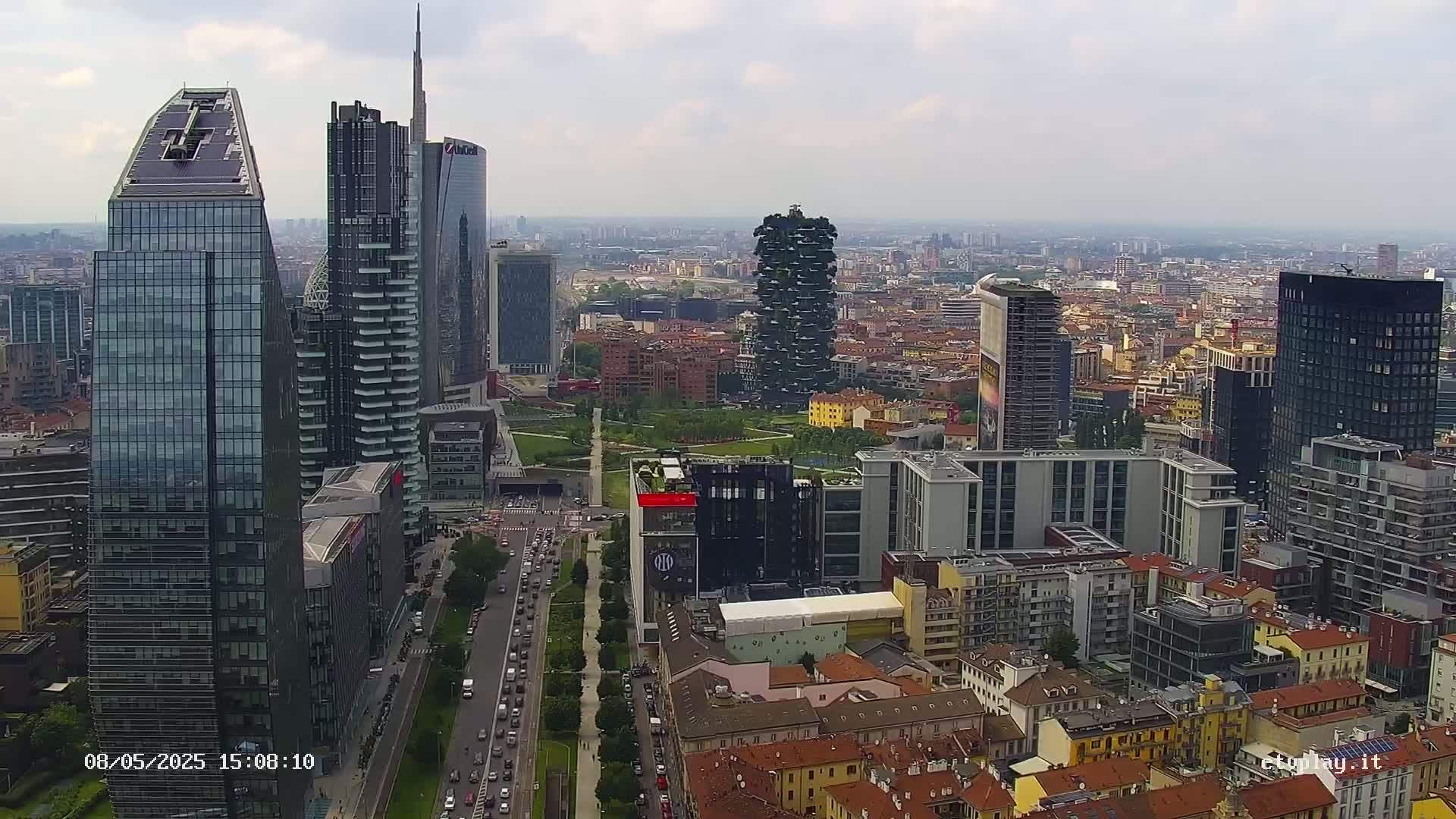 An aerial view shows a cityscape of numerous modern skyscrapers and traditional buildings under a cloudy sky.