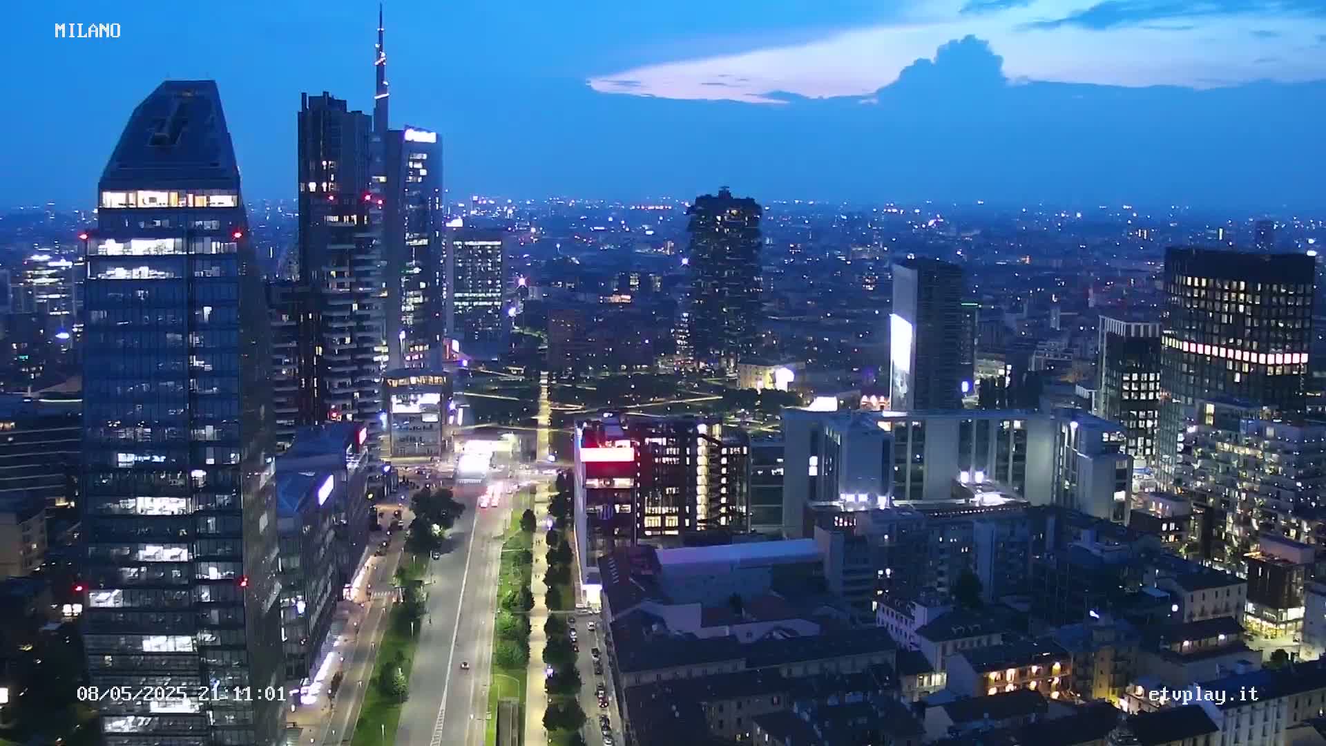 A nighttime aerial view of a city skyline with many illuminated skyscrapers and a long, straight road, under a twilight sky.