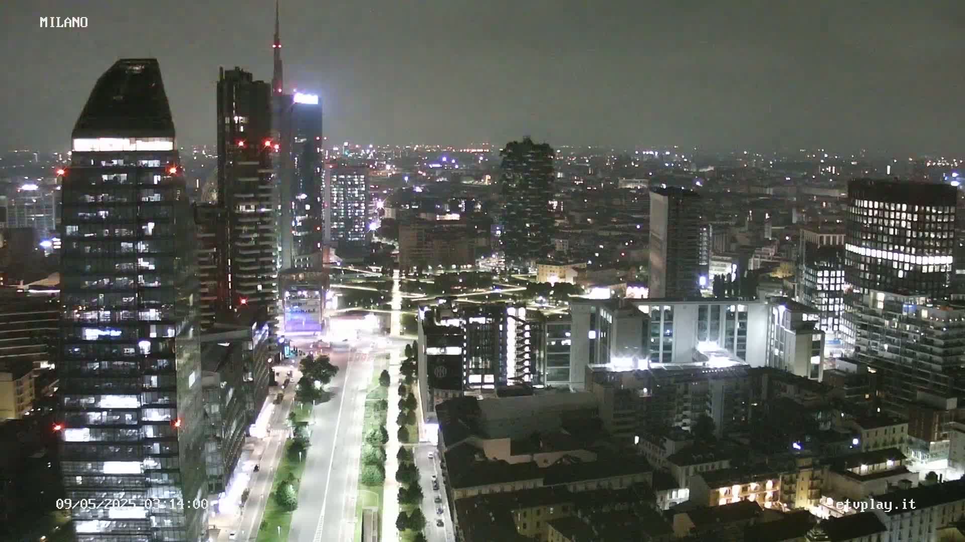 A nighttime aerial view of a city skyline showing many illuminated skyscrapers and buildings, under a dark, overcast sky.