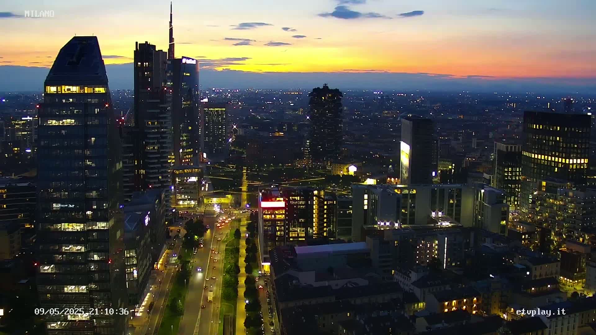 An aerial view shows a city skyline at twilight, with numerous skyscrapers illuminated against a colorful sunset sky.