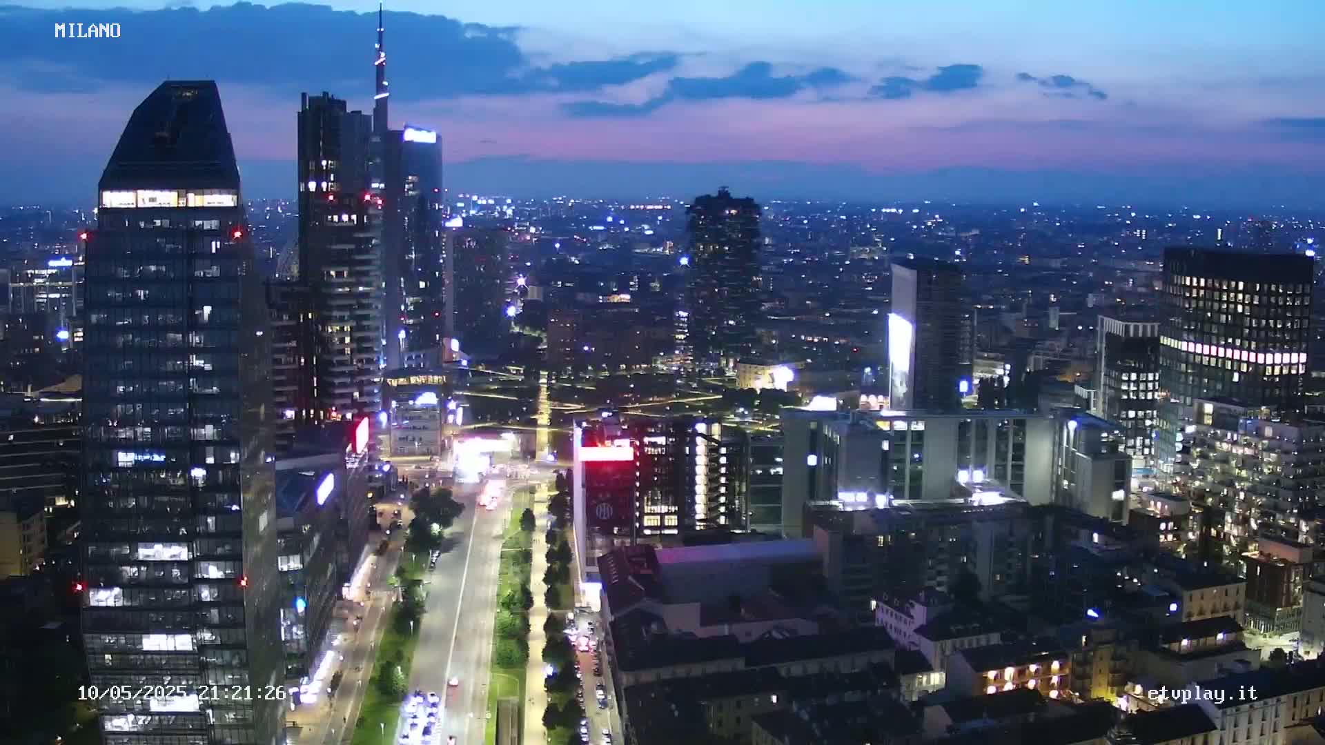 A nighttime aerial view of a city skyline shows numerous illuminated skyscrapers and buildings under a twilight sky.