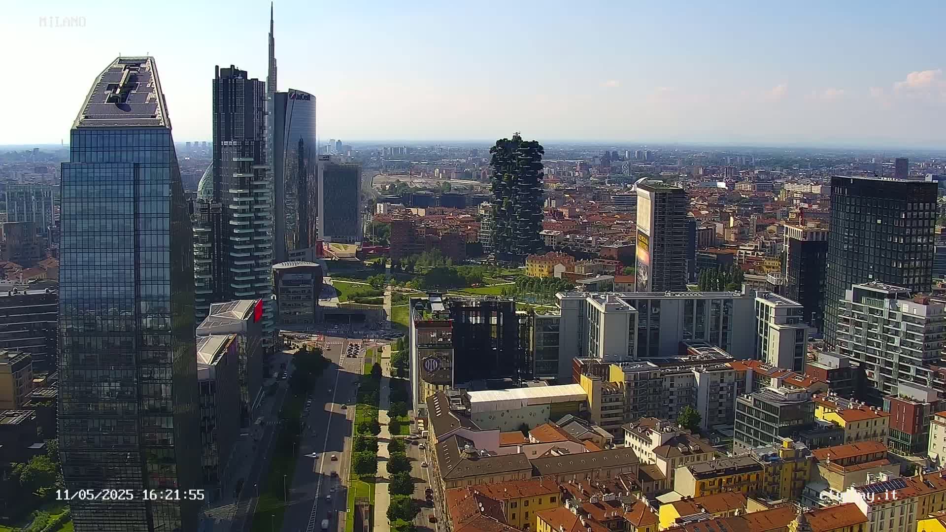 A high-angle, sunny view of a cityscape featuring numerous modern skyscrapers and surrounding low-rise buildings, with a park visible between the taller structures.