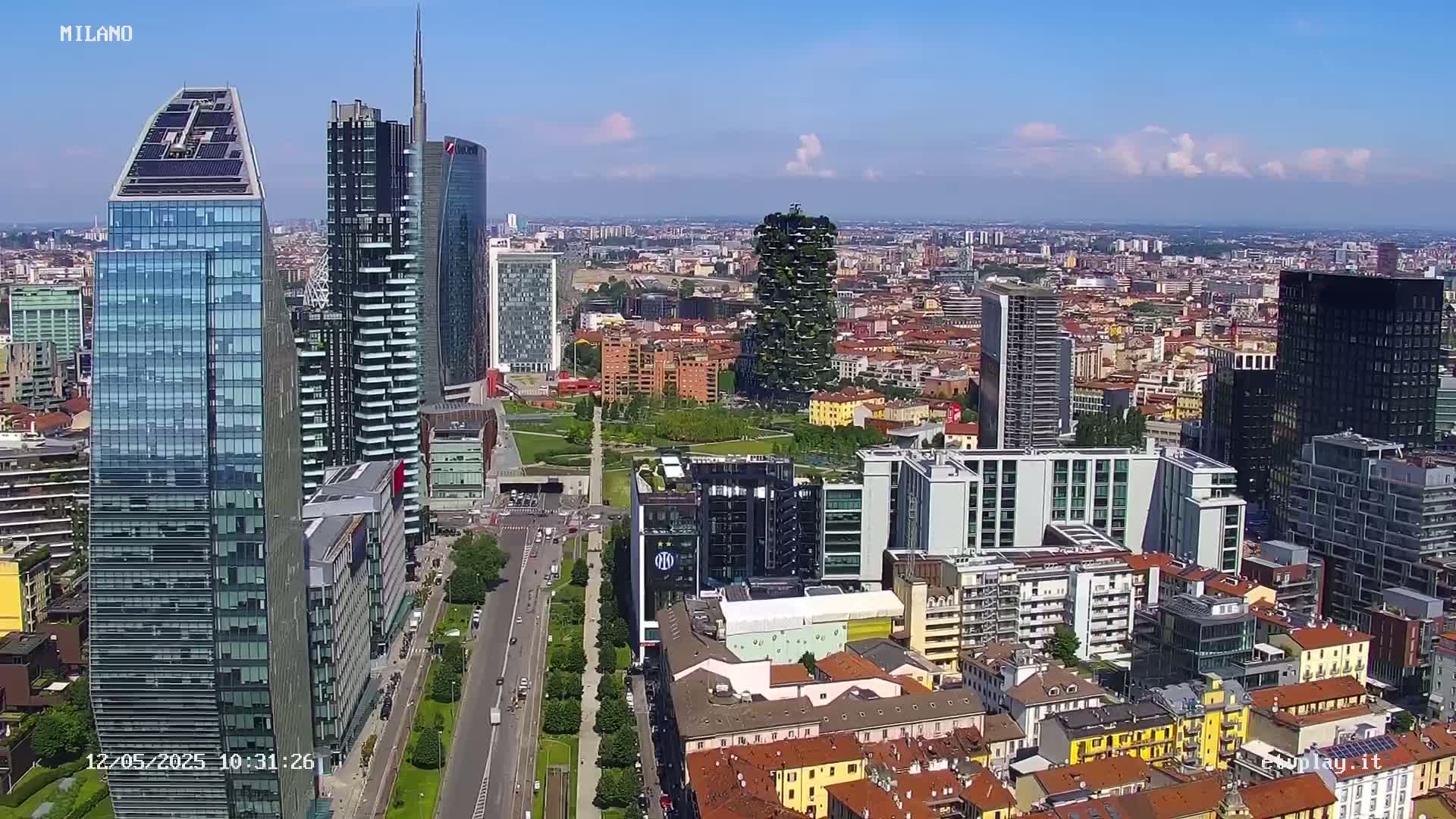 A high-angle, sunny-day view of a dense cityscape featuring numerous modern skyscrapers and surrounding low-rise buildings, with a large park visible in the center.