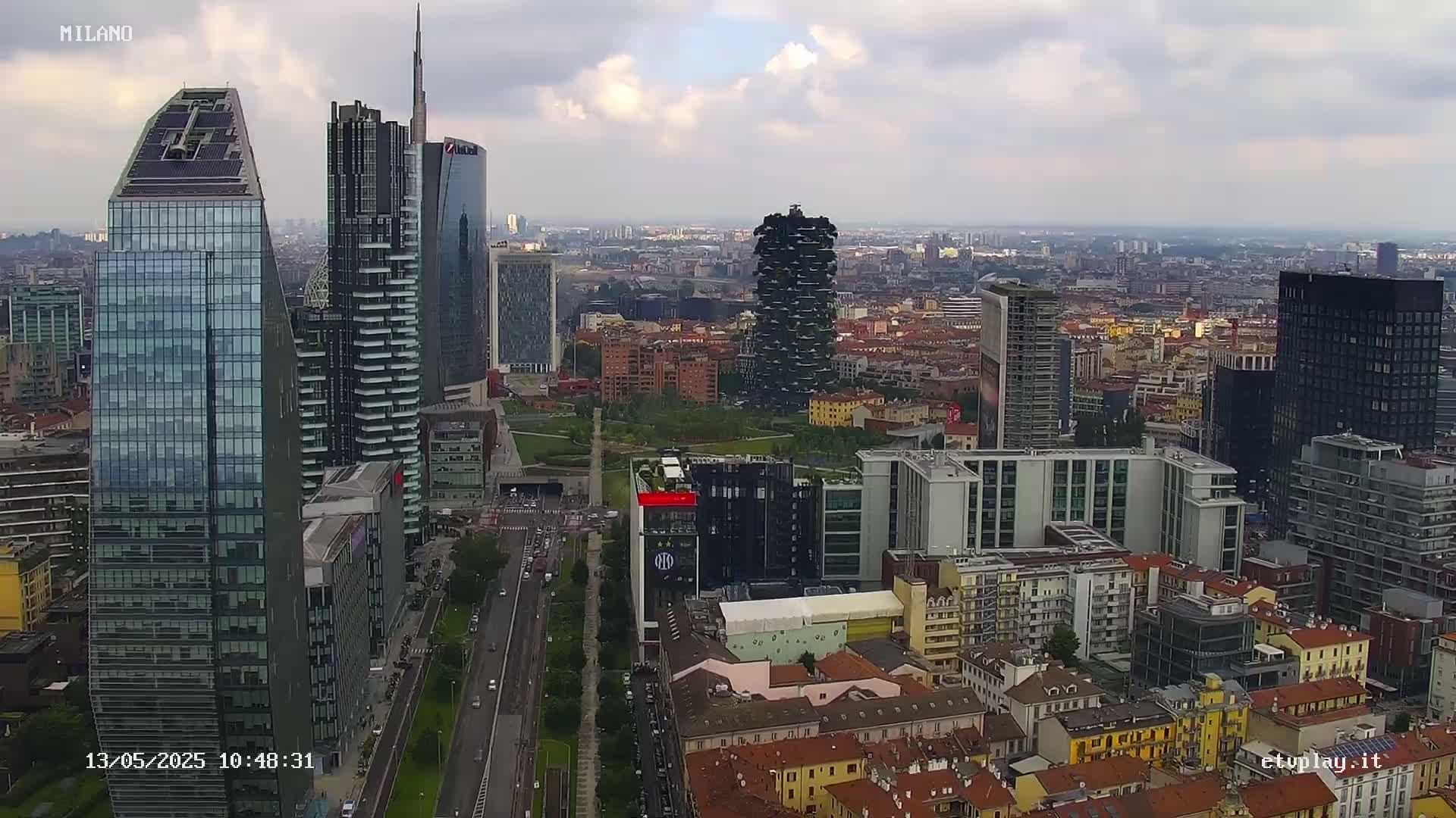 An aerial view of a city, under a cloudy sky, shows a mix of modern skyscrapers and low-rise buildings.
