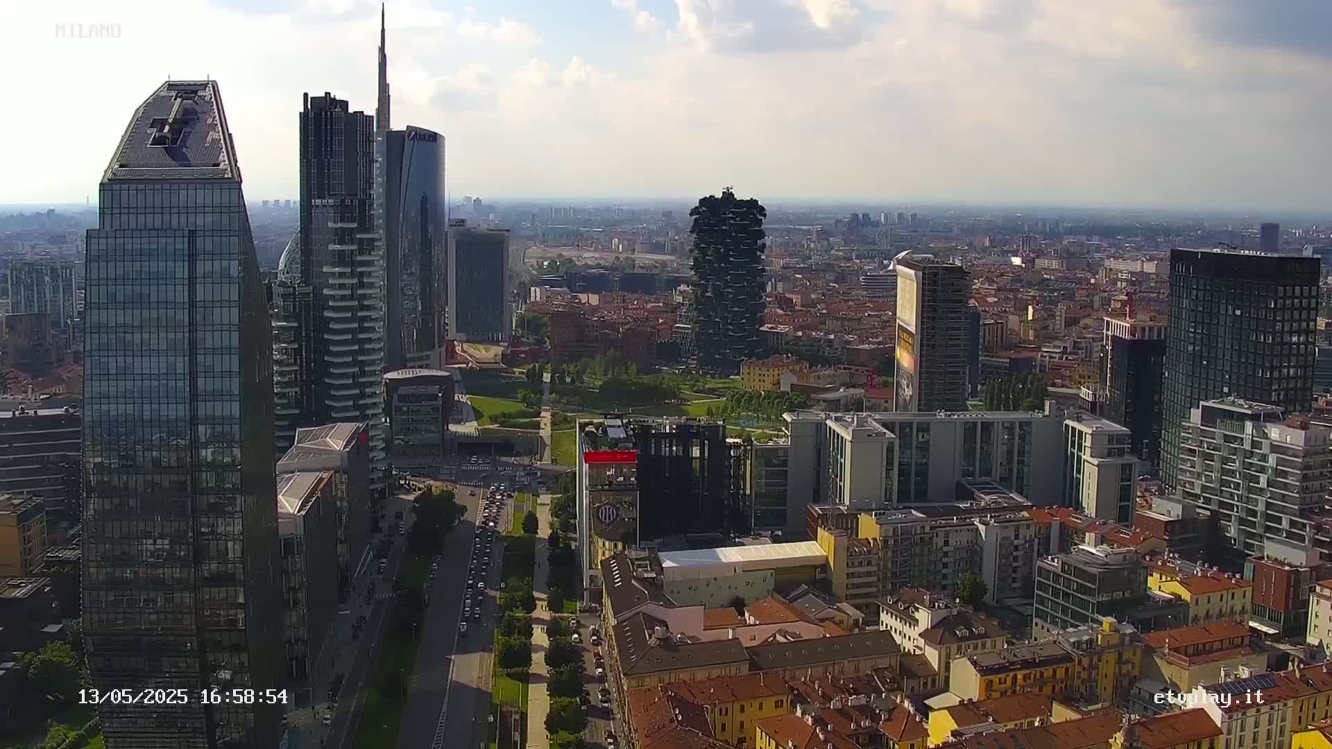 An aerial view of a city with many modern skyscrapers and surrounding low-rise buildings on a partly cloudy day.