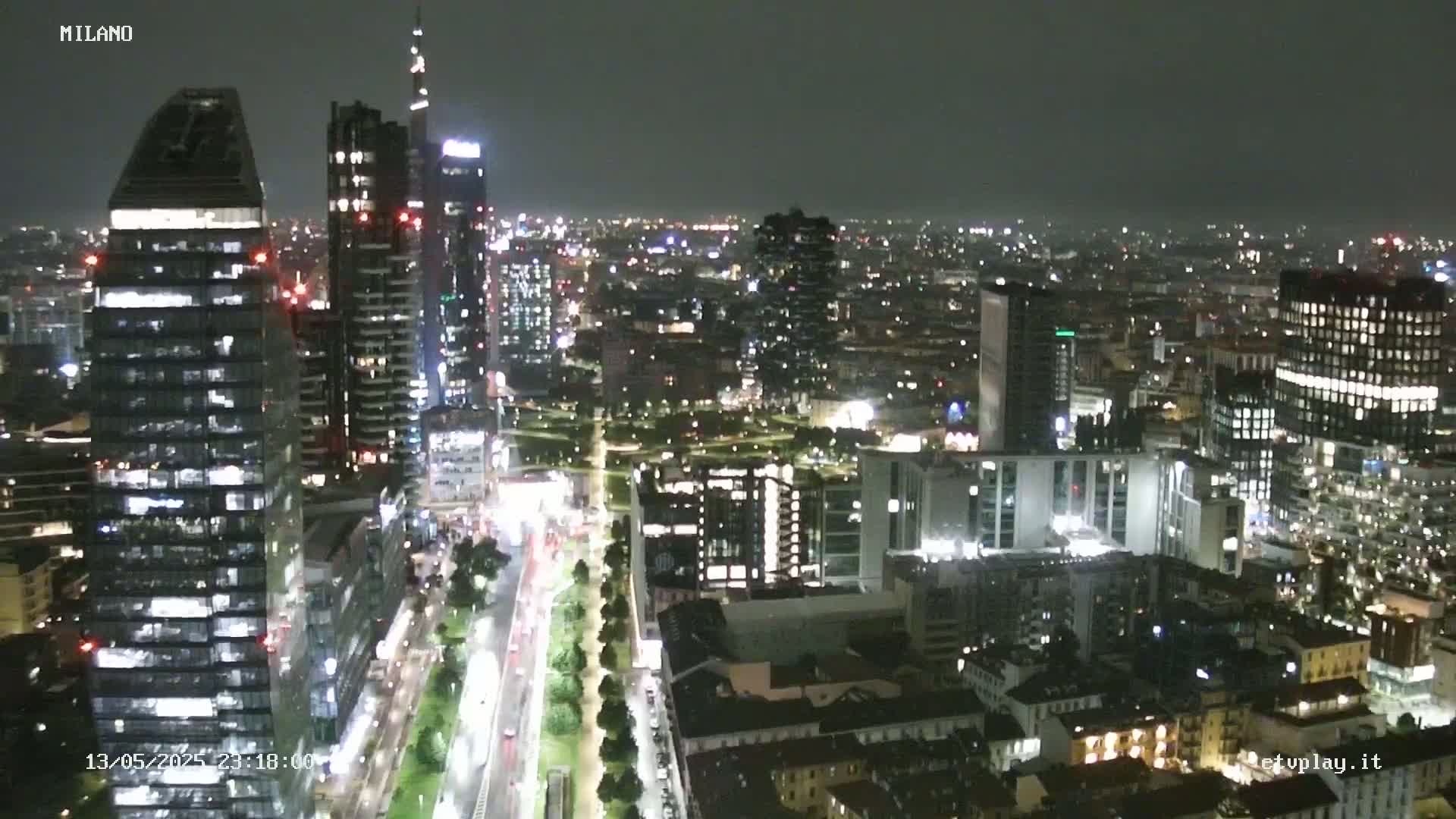 A nighttime aerial view of a city, showing brightly lit skyscrapers and streets with moving vehicles under a dark sky.