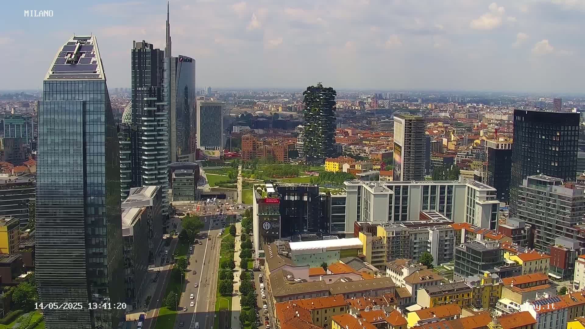 A high-angle, sunny view shows a city skyline with numerous skyscrapers and a park, and a long, wide road running through the city.