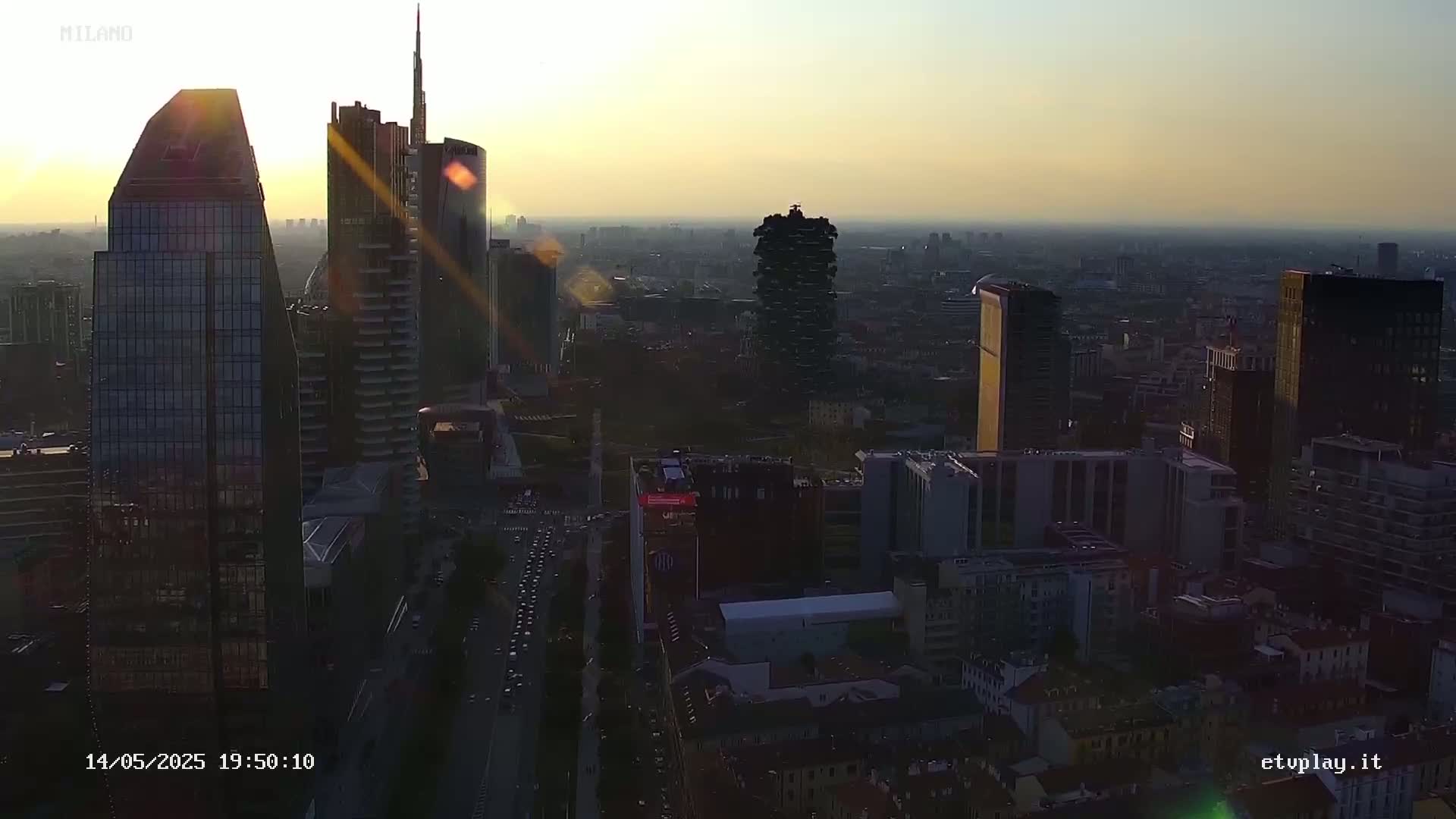 A hazy sunset illuminates a cityscape of tall, modern buildings and a network of streets with traffic, seen from a high vantage point.