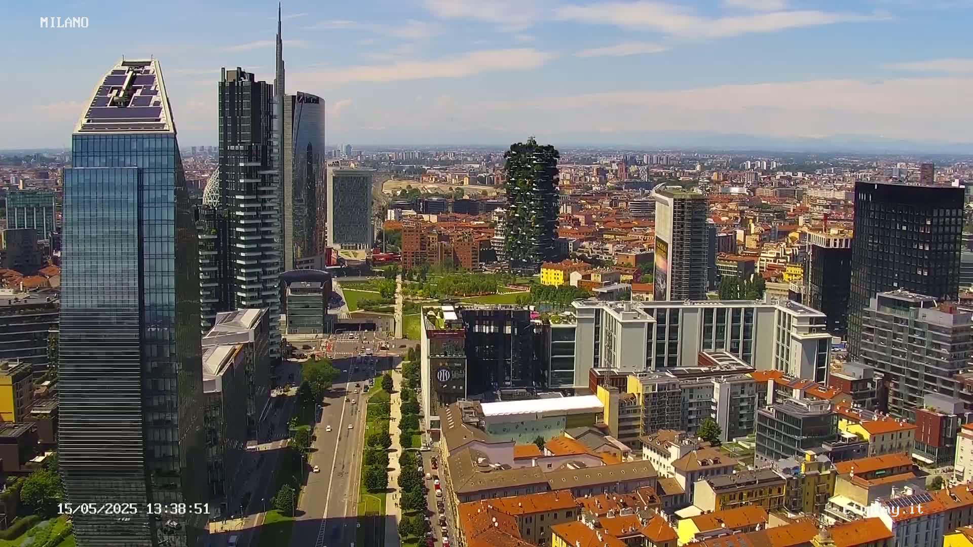 An aerial view of a city on a sunny day shows many modern skyscrapers and smaller buildings, with a large park visible between some of the taller structures.