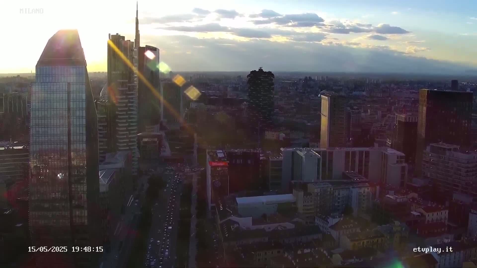 An aerial view of a city skyline at sunset, with skyscrapers and lens flare from the setting sun.