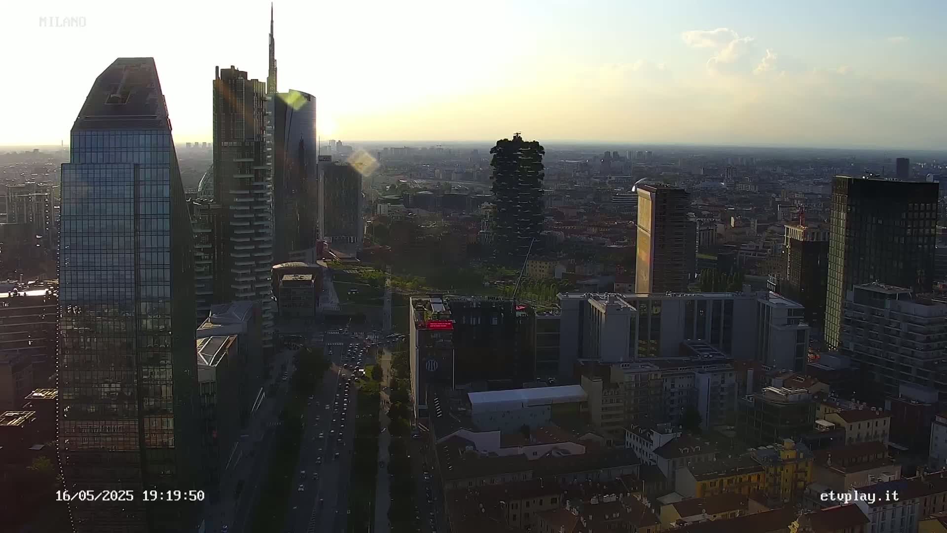 An aerial view of a dense cityscape at sunset, showing numerous skyscrapers and surrounding buildings, with the sun partially obscured by some clouds.