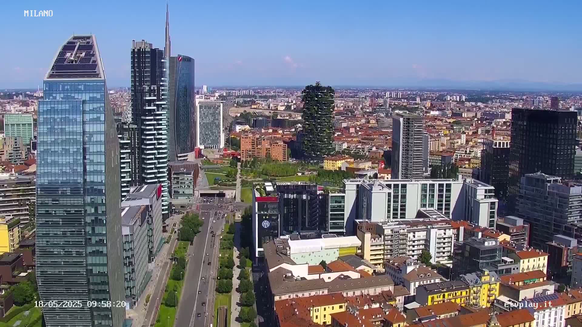 A high-angle, long shot of a city skyline on a clear, sunny day, showing numerous skyscrapers and surrounding buildings.