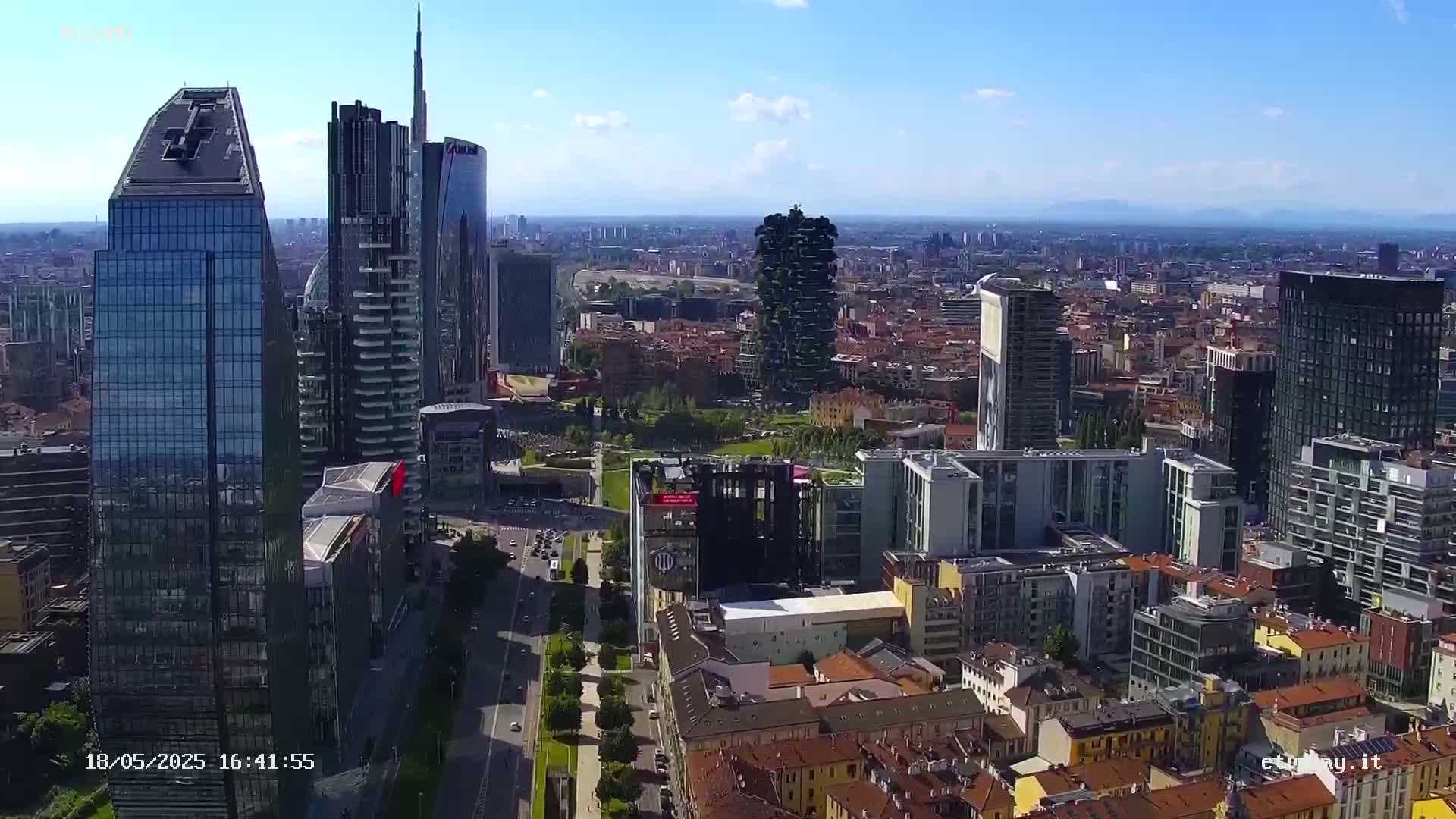 An aerial view of a city on a clear, sunny day, showing a mix of modern skyscrapers and traditional lower buildings.