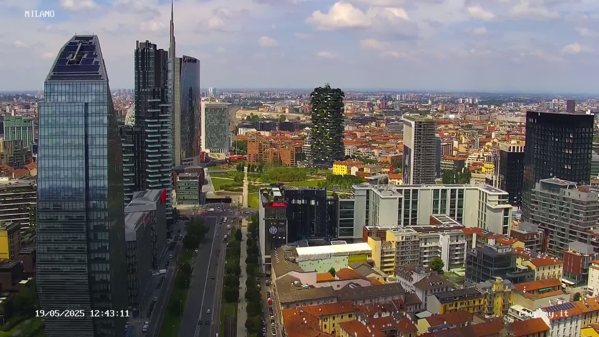 An aerial view of a city's skyline on a partly cloudy day, showing a mix of modern skyscrapers and older buildings, with a large green park visible in the mid-ground.
