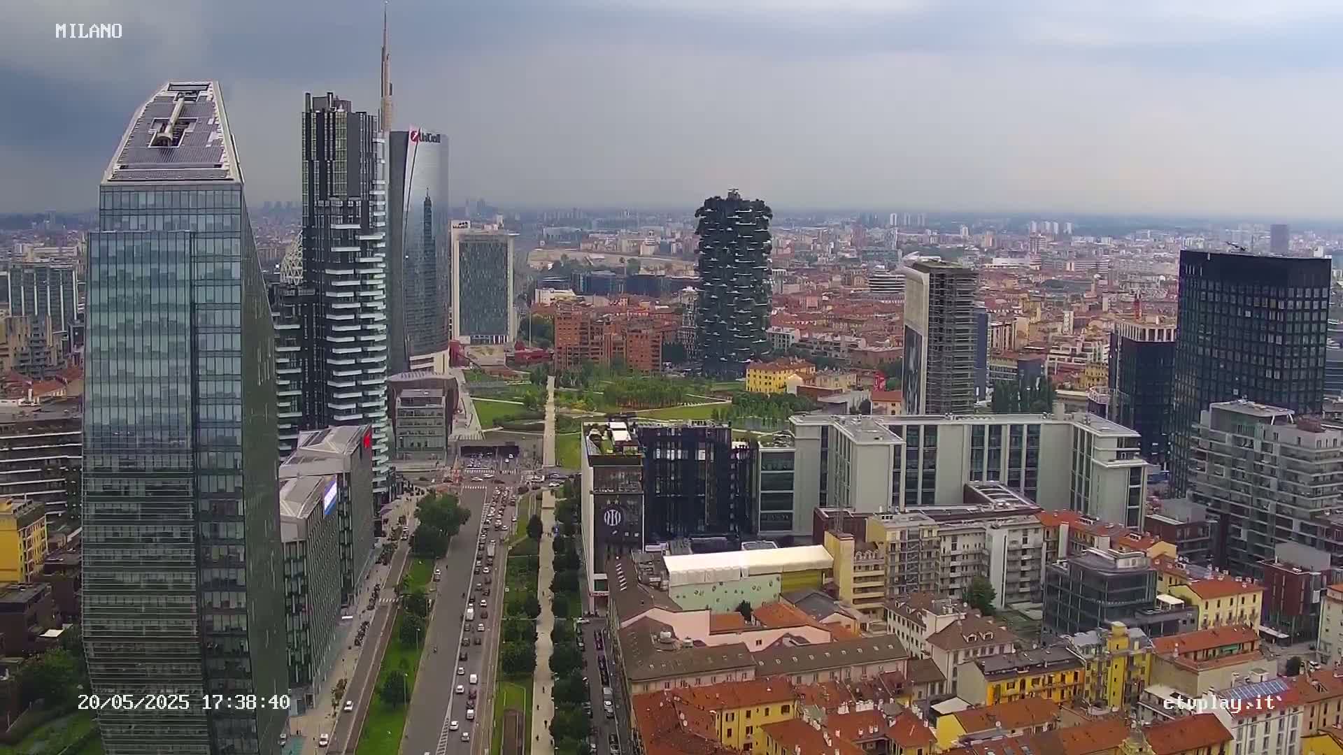 An aerial view of a city, under an overcast sky, shows numerous skyscrapers and high-rise buildings surrounding a large park.