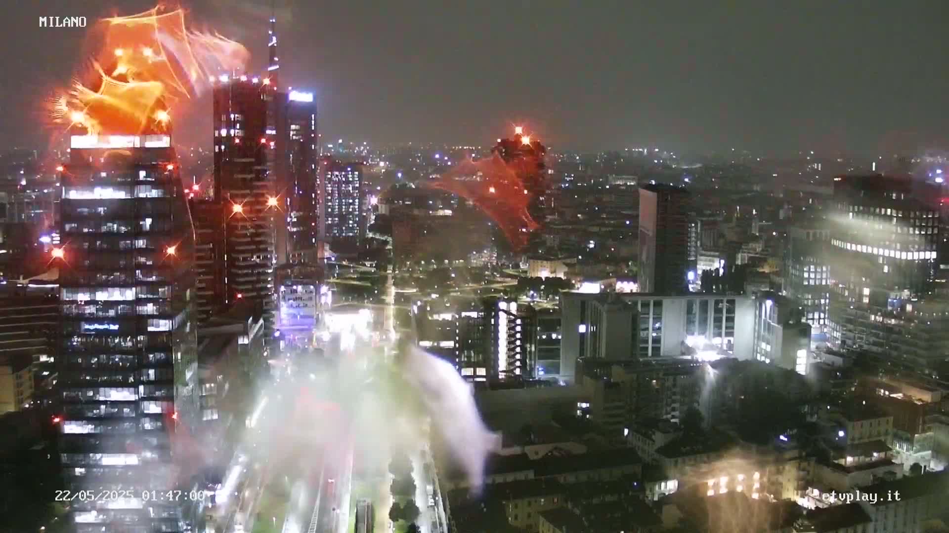 Orange fireworks explode over a cityscape at night, illuminating numerous buildings.