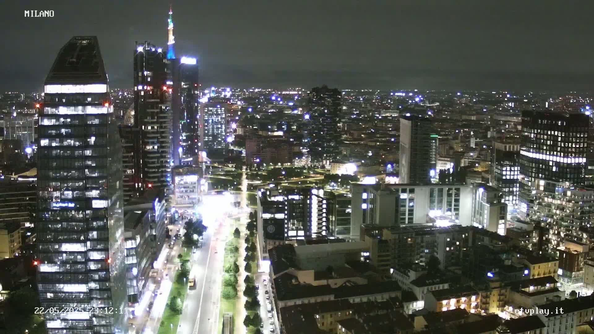 A nighttime aerial view of a city's skyline, showing numerous illuminated buildings and streets with cars driving on them.