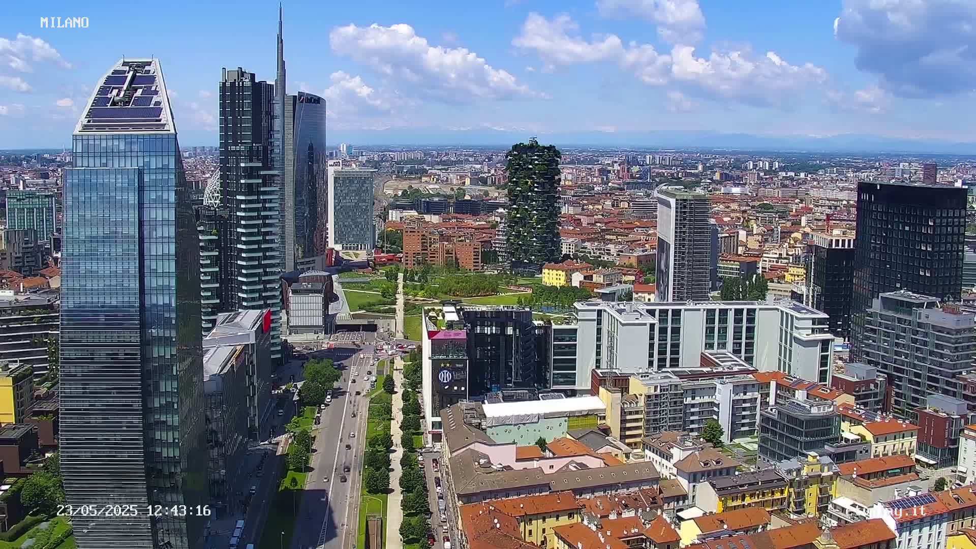 A high-angle, sunny view of a dense cityscape with numerous skyscrapers and a large park, under a partly cloudy sky.