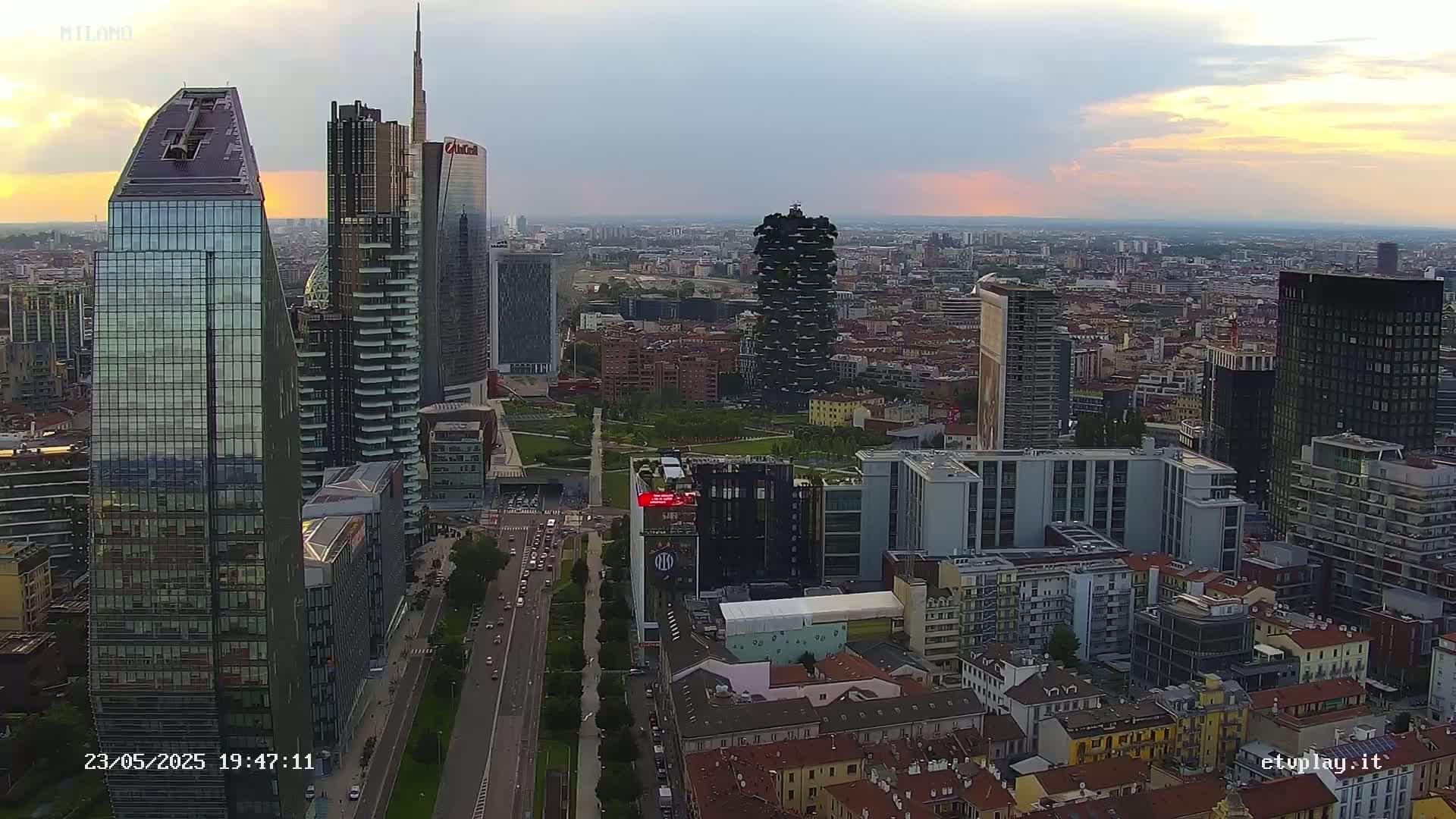 An aerial view of a city skyline at sunset, showing numerous skyscrapers and surrounding buildings under partly cloudy skies.