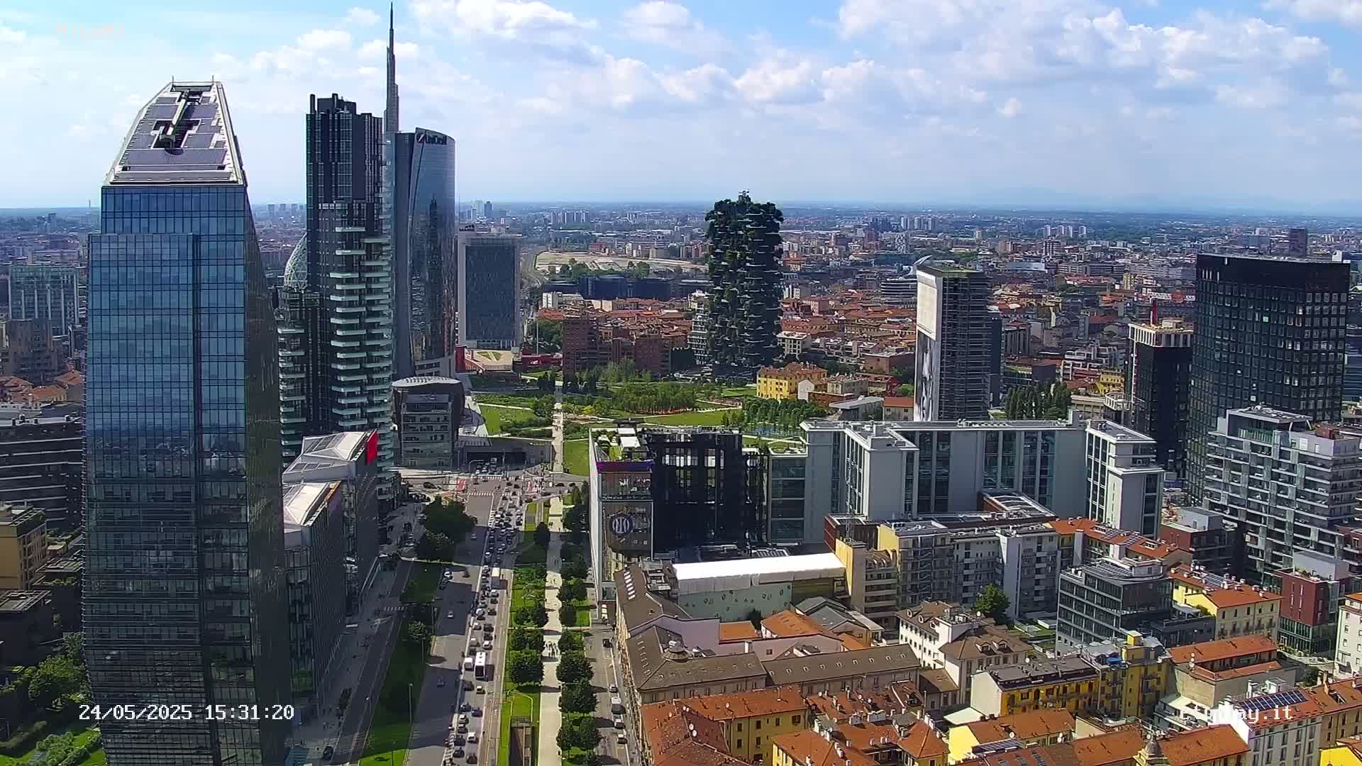 An aerial view of a city on a partly cloudy day shows numerous skyscrapers and surrounding low-rise buildings, with a park visible between some of the taller structures.