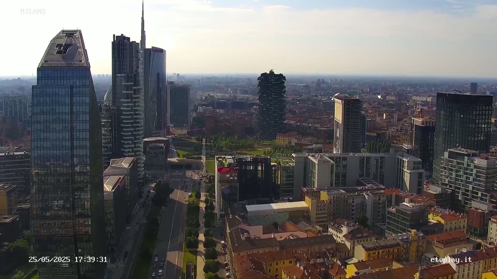 An aerial view of a city on a partly sunny day shows a mix of modern skyscrapers and older, low-rise buildings.