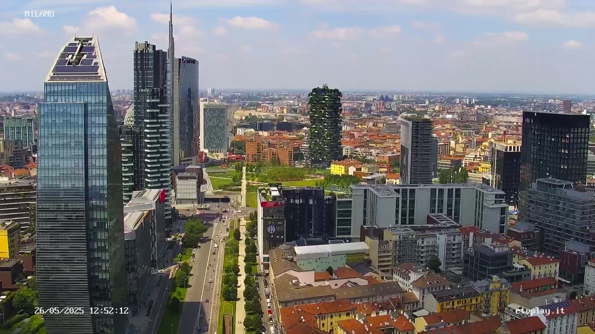 An aerial view of a city skyline on a partly cloudy day shows numerous skyscrapers and surrounding buildings, with a park and roads visible.