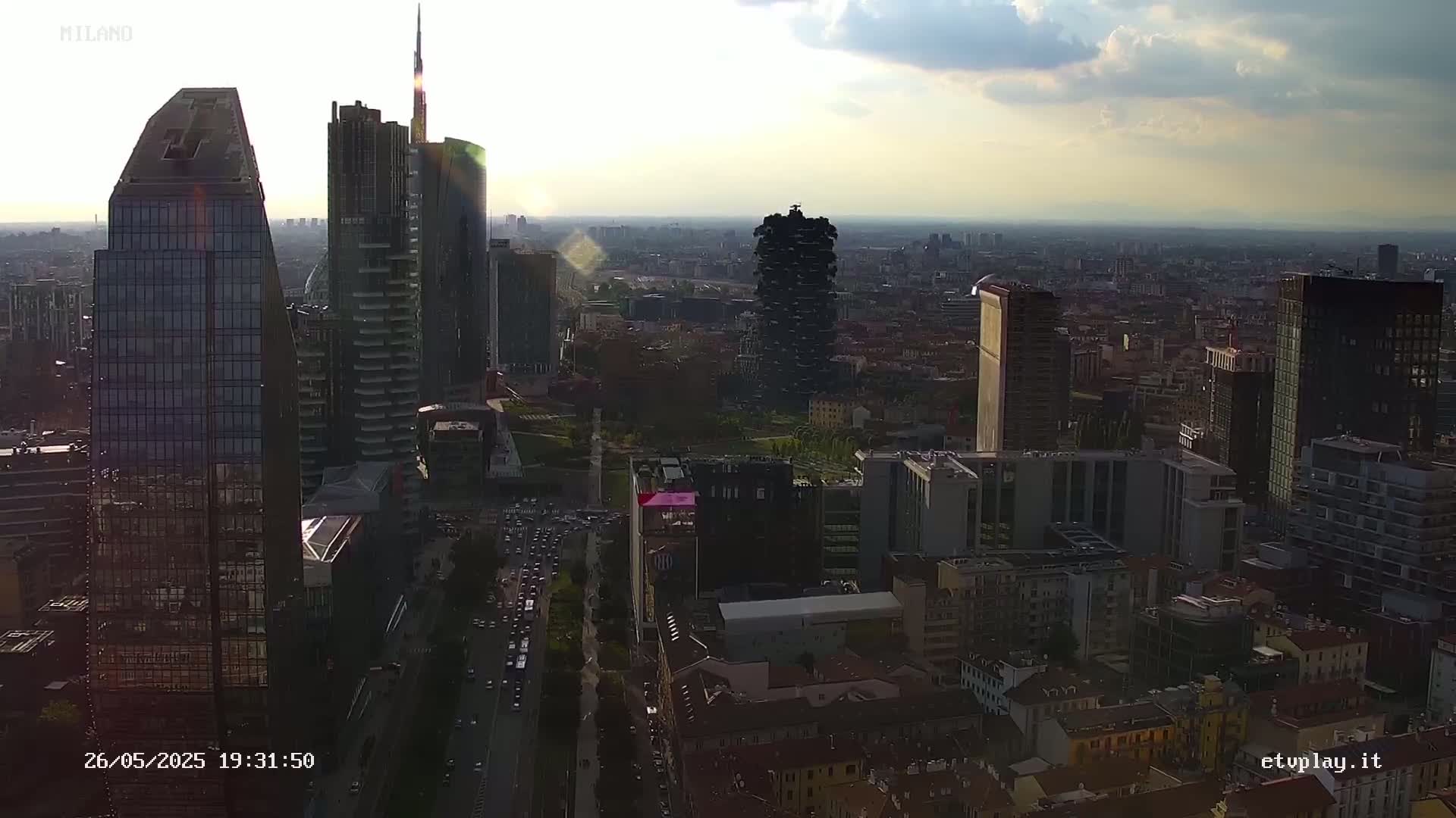 An aerial view of a city skyline under a partly cloudy sky shows numerous skyscrapers and surrounding buildings at sunset.