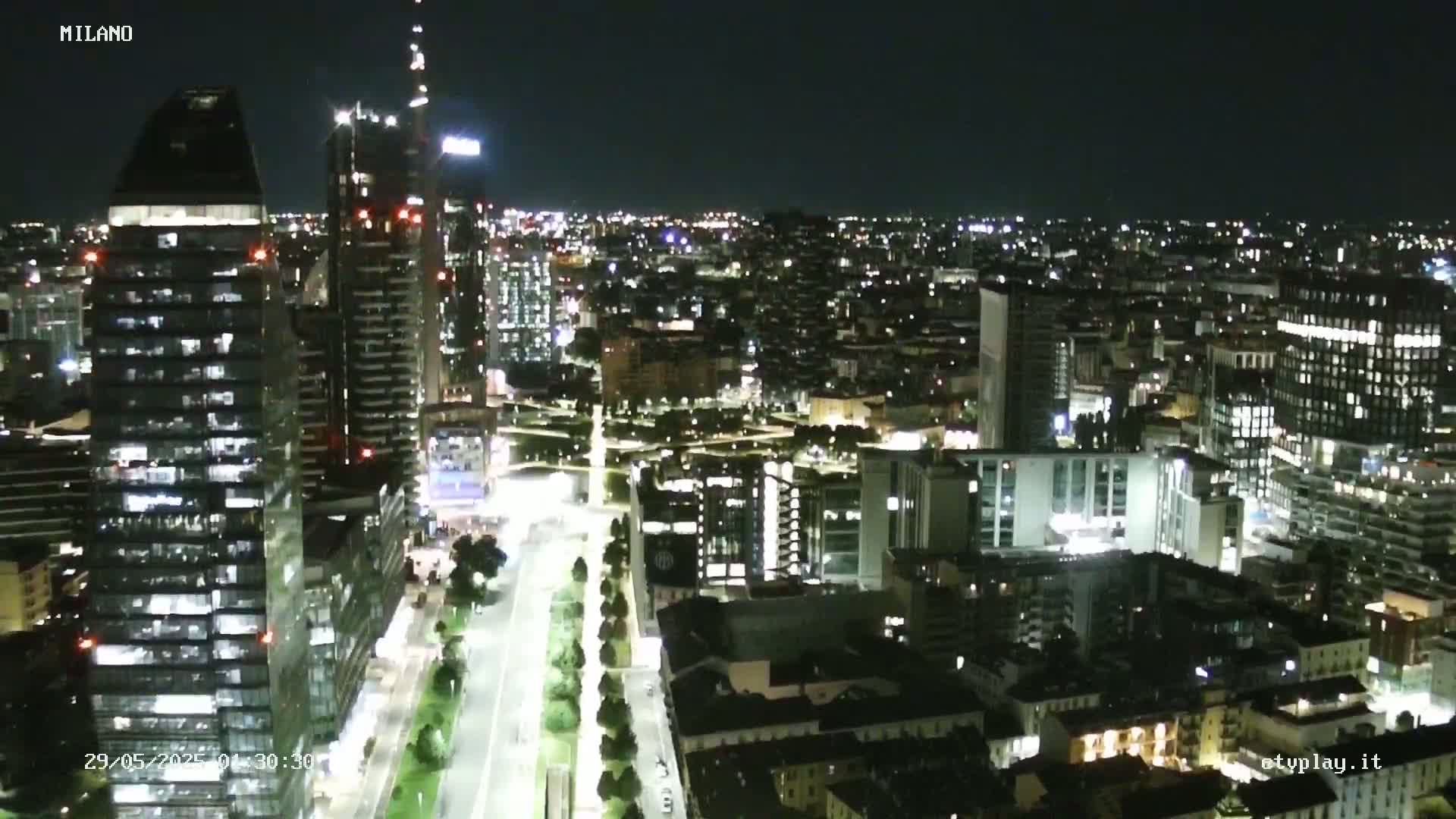 A nighttime aerial view of a city with numerous illuminated buildings and a long, tree-lined road.