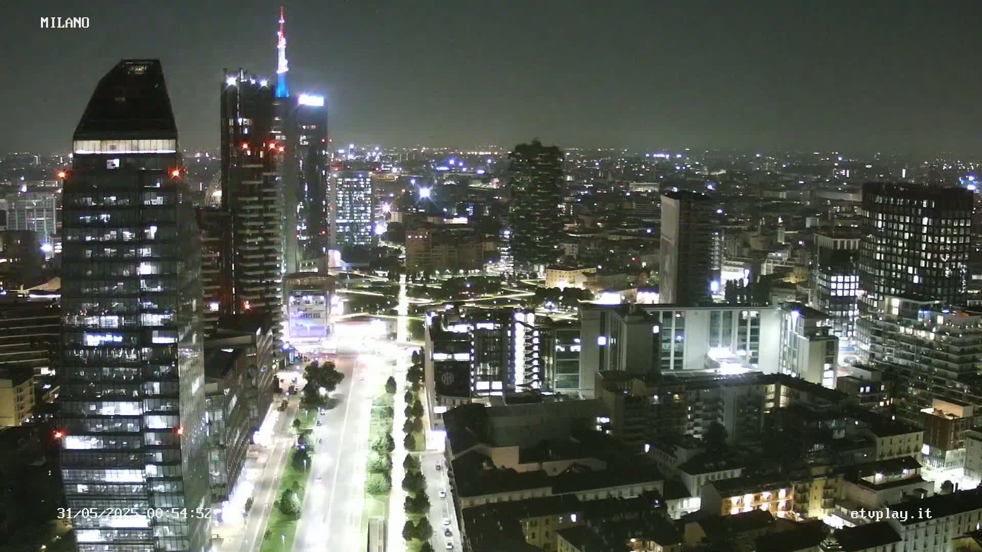A nighttime aerial view of a city with many tall buildings illuminated, showing a clear sky.