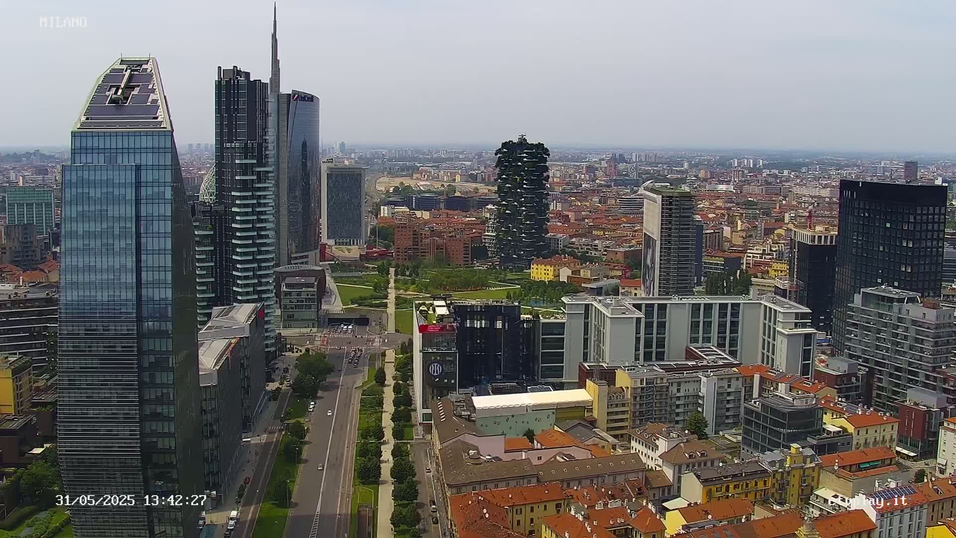 An aerial view of a city on a clear day shows a mix of modern skyscrapers and older, lower buildings, with a park separating some of the taller structures.
