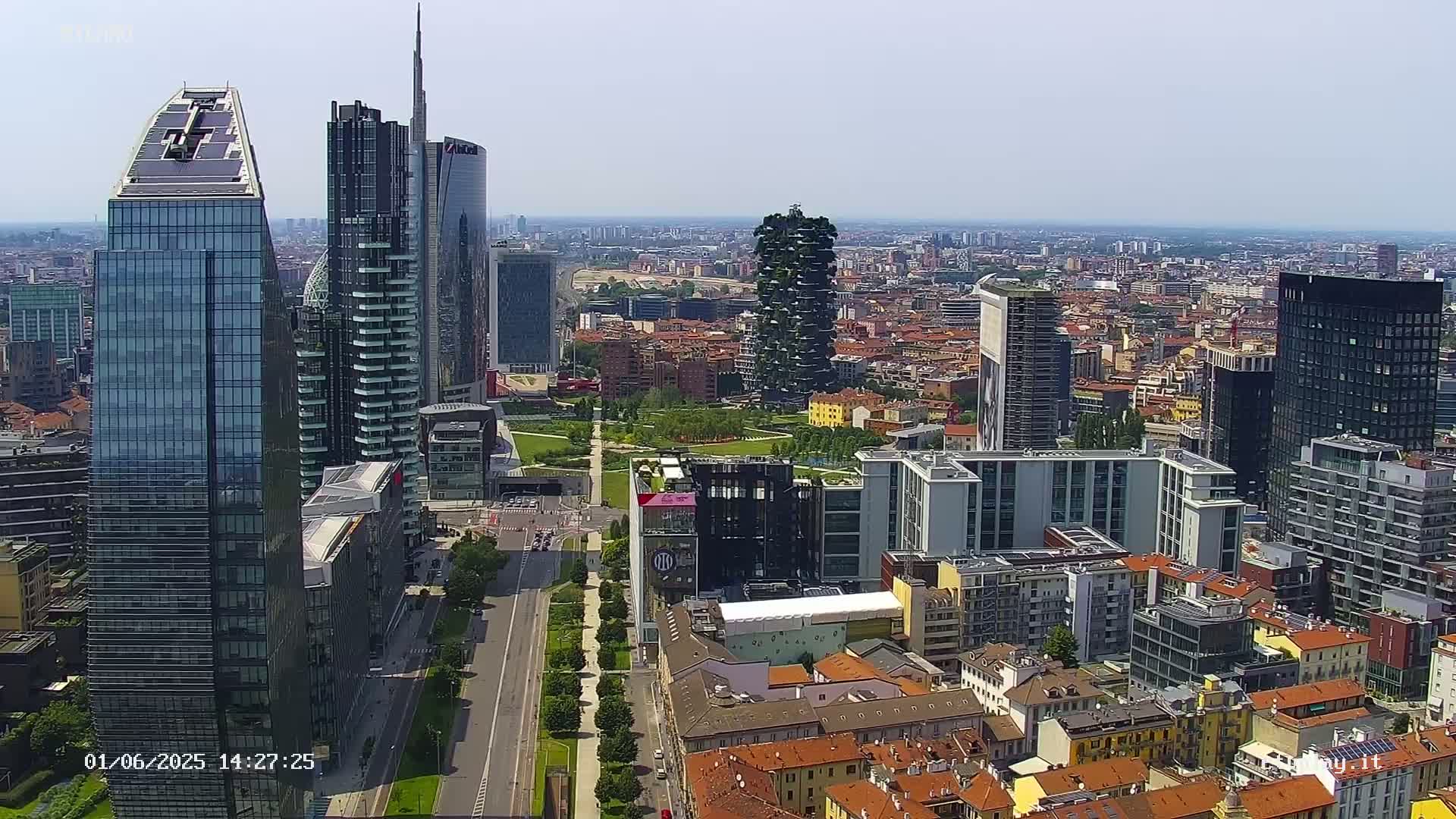 An aerial view of a city on a sunny day shows a mix of modern skyscrapers and older, low-rise buildings, with a large park separating some of the taller structures.