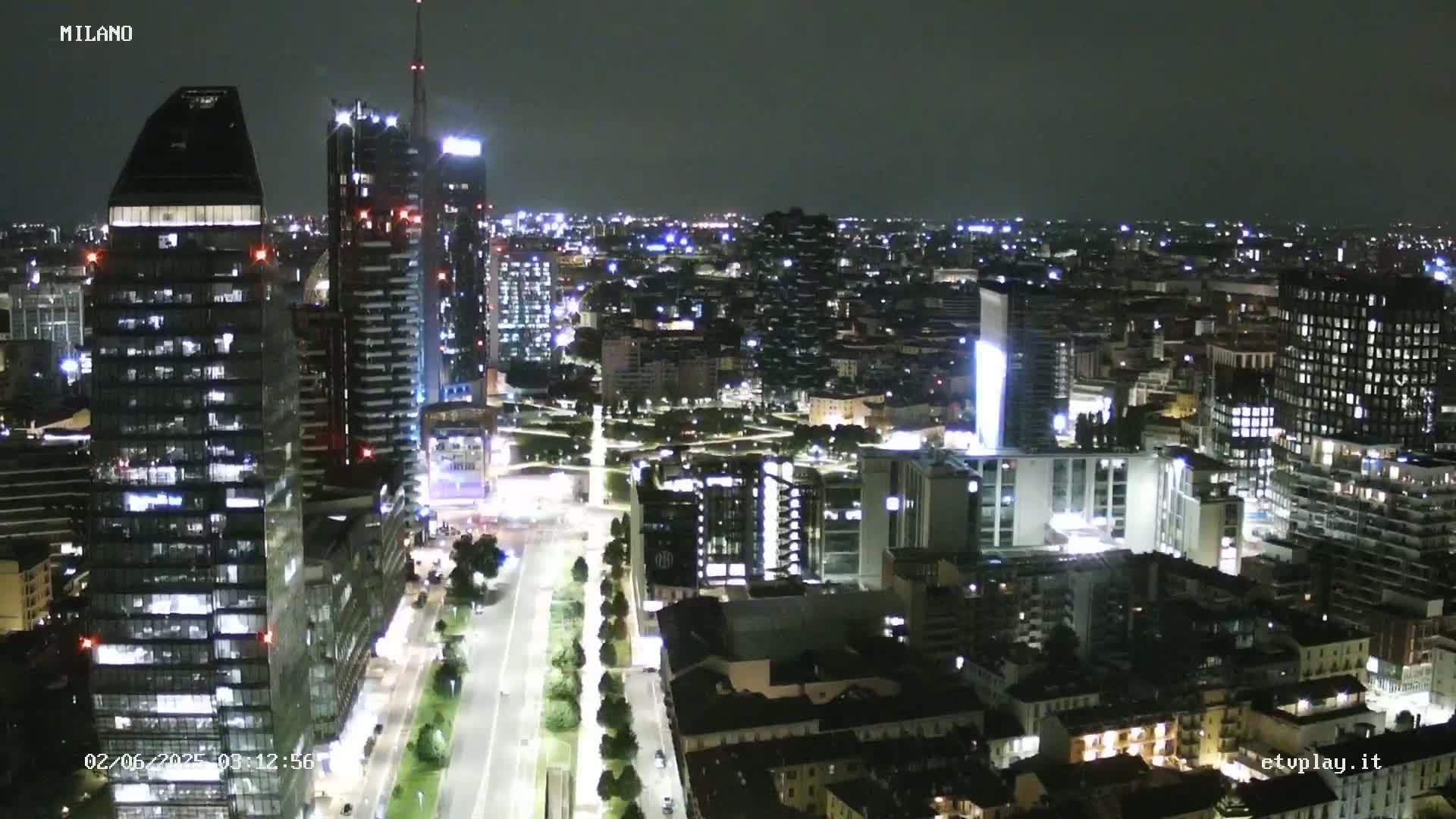 A nighttime aerial view of a city's skyline, showing brightly lit skyscrapers and streets, under a dark sky.