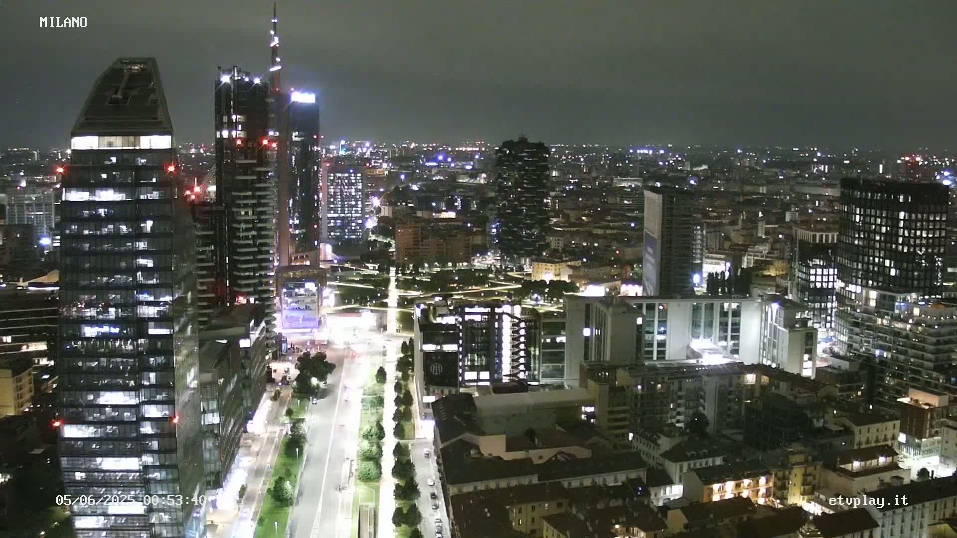 A nighttime aerial view of a city with many tall buildings illuminated, showing a wide street with trees lining the sides and a park in the middle.