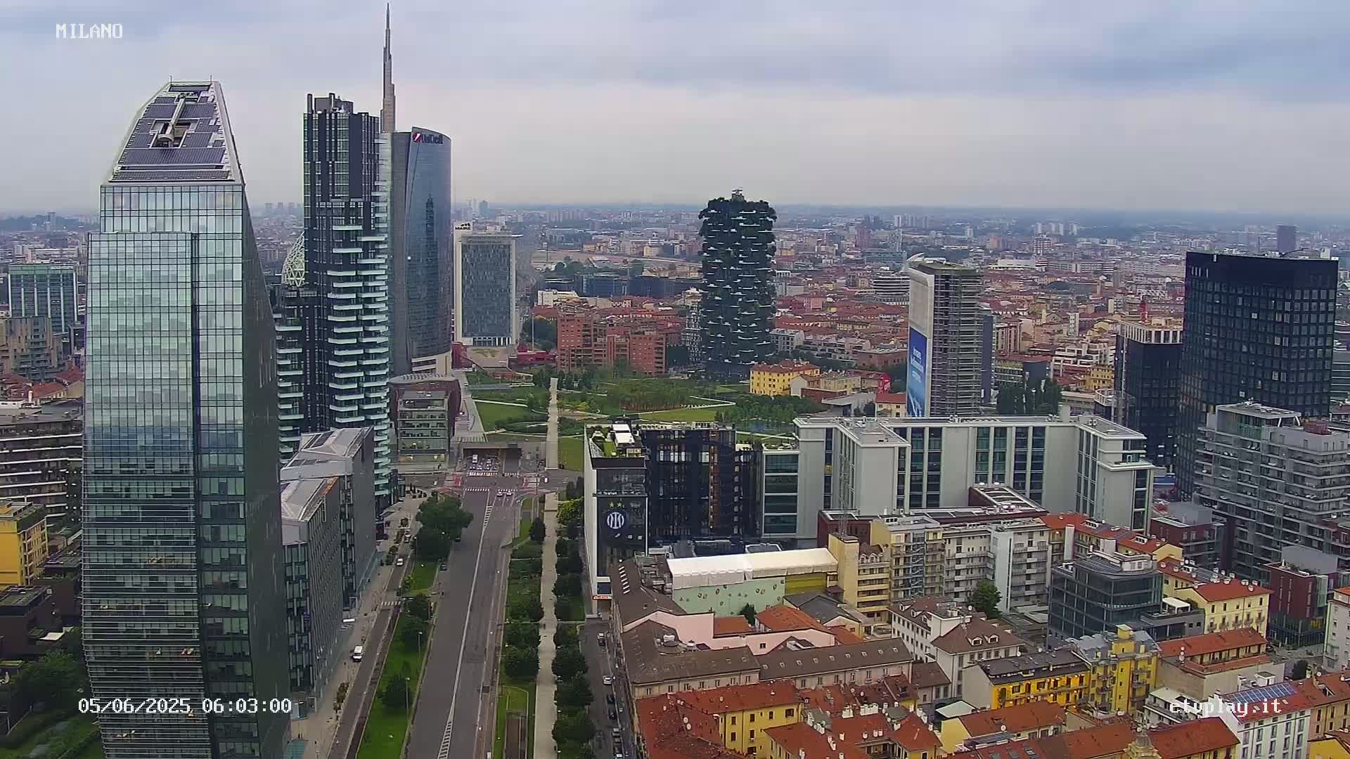 A high-angle, long shot of a city skyline on an overcast day, featuring numerous skyscrapers and surrounding buildings, with a large park visible in the mid-ground.