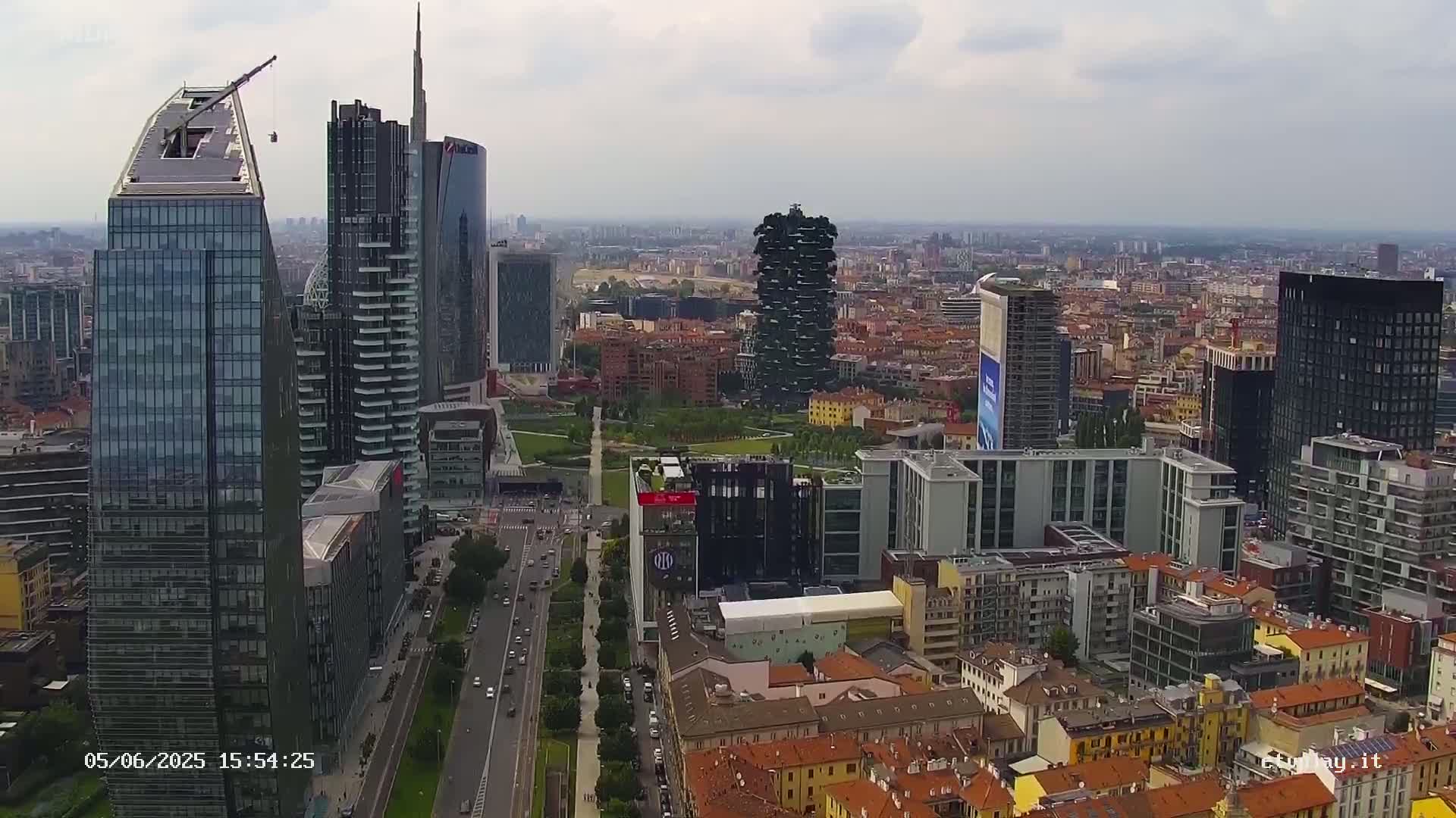 An aerial view of a city skyline shows numerous modern skyscrapers and other buildings under a mostly cloudy sky.
