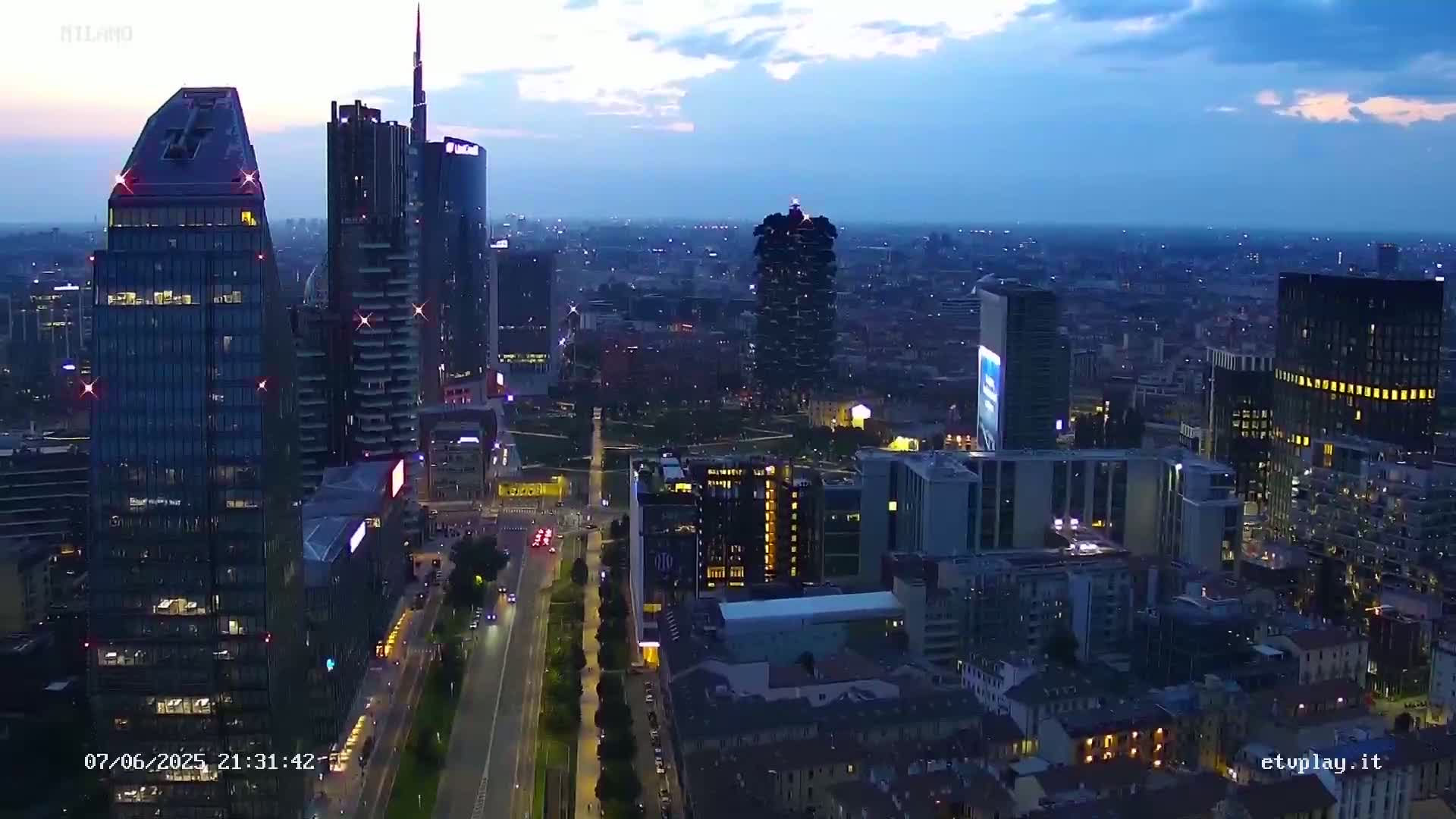 A high-angle, twilight view of a city skyline with numerous illuminated skyscrapers and a roadway cutting through a cityscape.