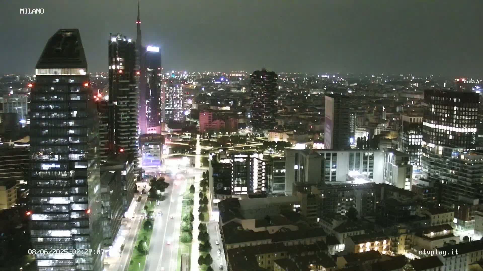 A nighttime aerial view of a city with numerous skyscrapers and brightly lit streets under a dark sky.