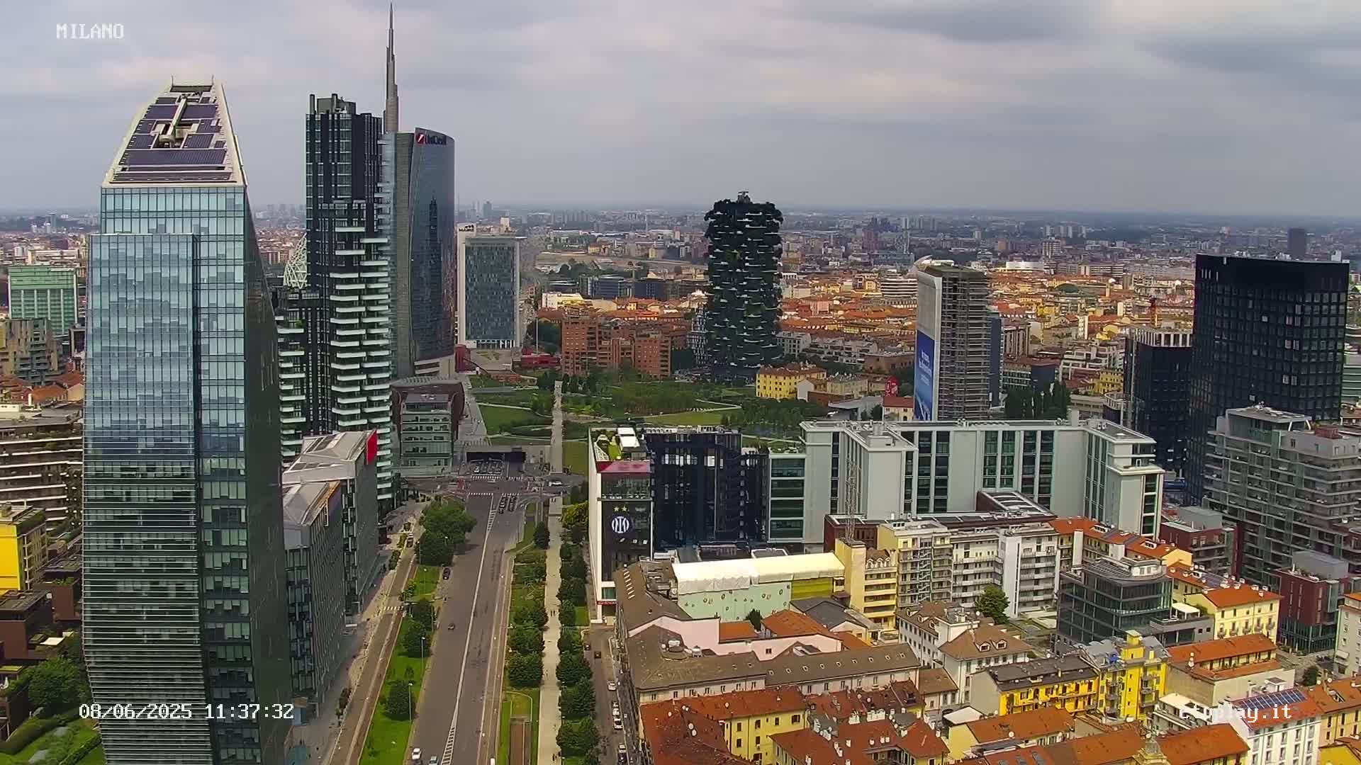 An aerial view of a city with numerous skyscrapers under cloudy skies shows a wide, tree-lined road cutting through the city.