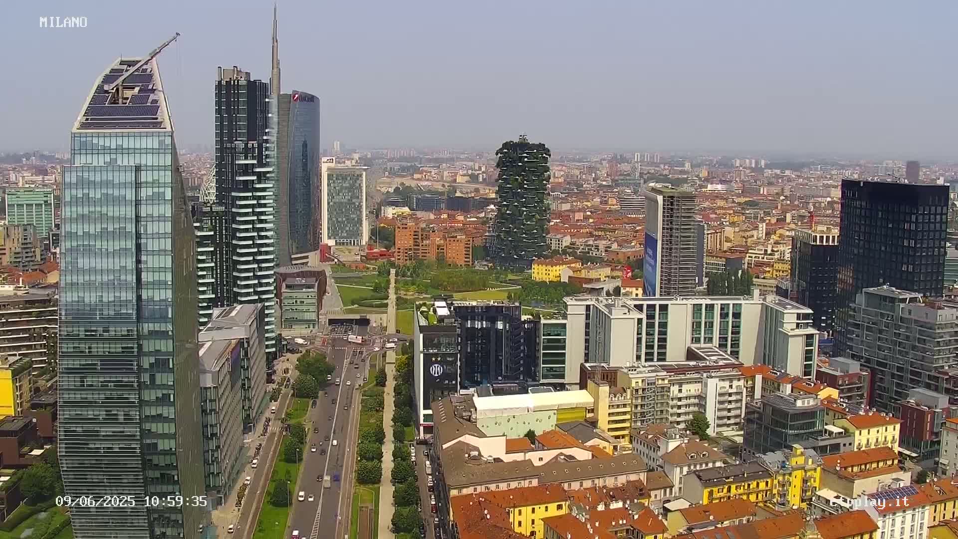 A high-angle, long shot of a city skyline on a clear, sunny day shows many skyscrapers and high-rise buildings, along with tree-lined streets and a large park.