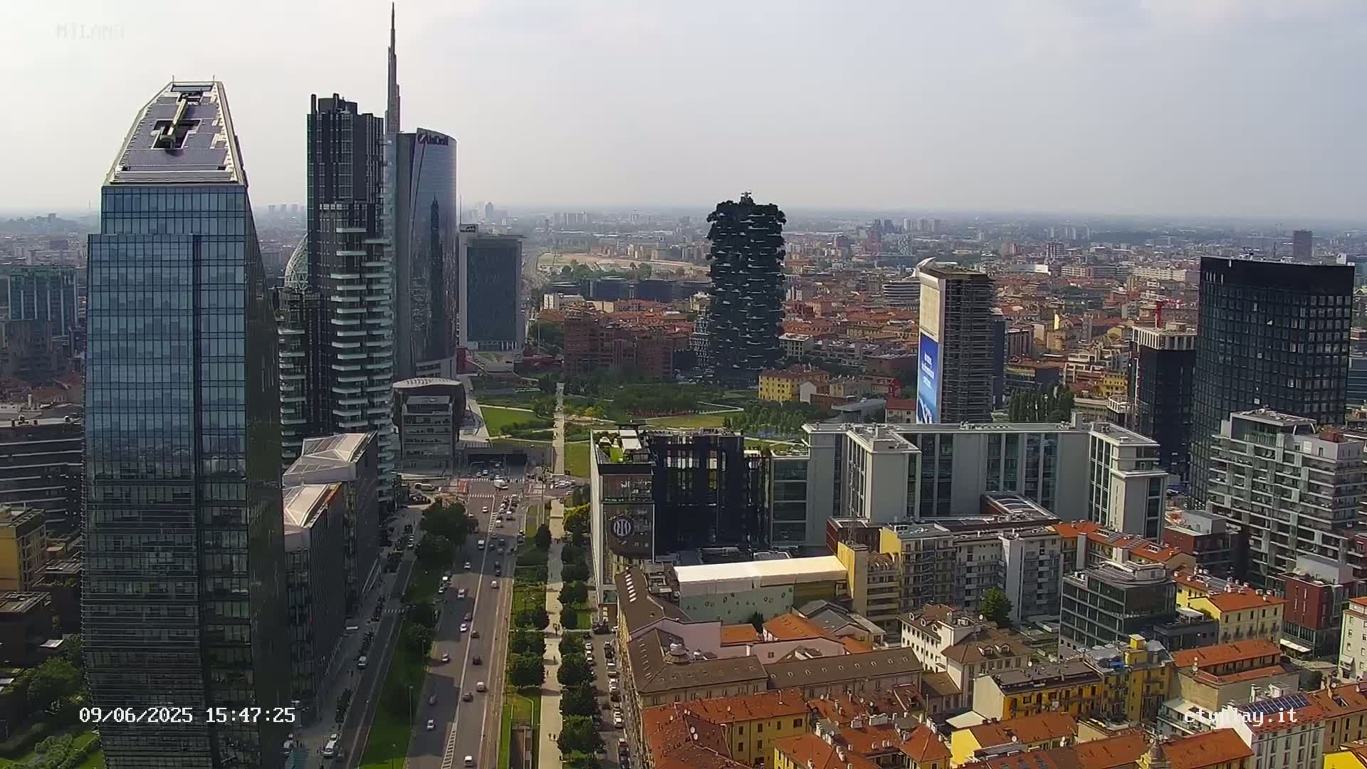 A high-angle, long shot shows a cityscape of modern and traditional buildings under a hazy sky, with a city park visible in the center.